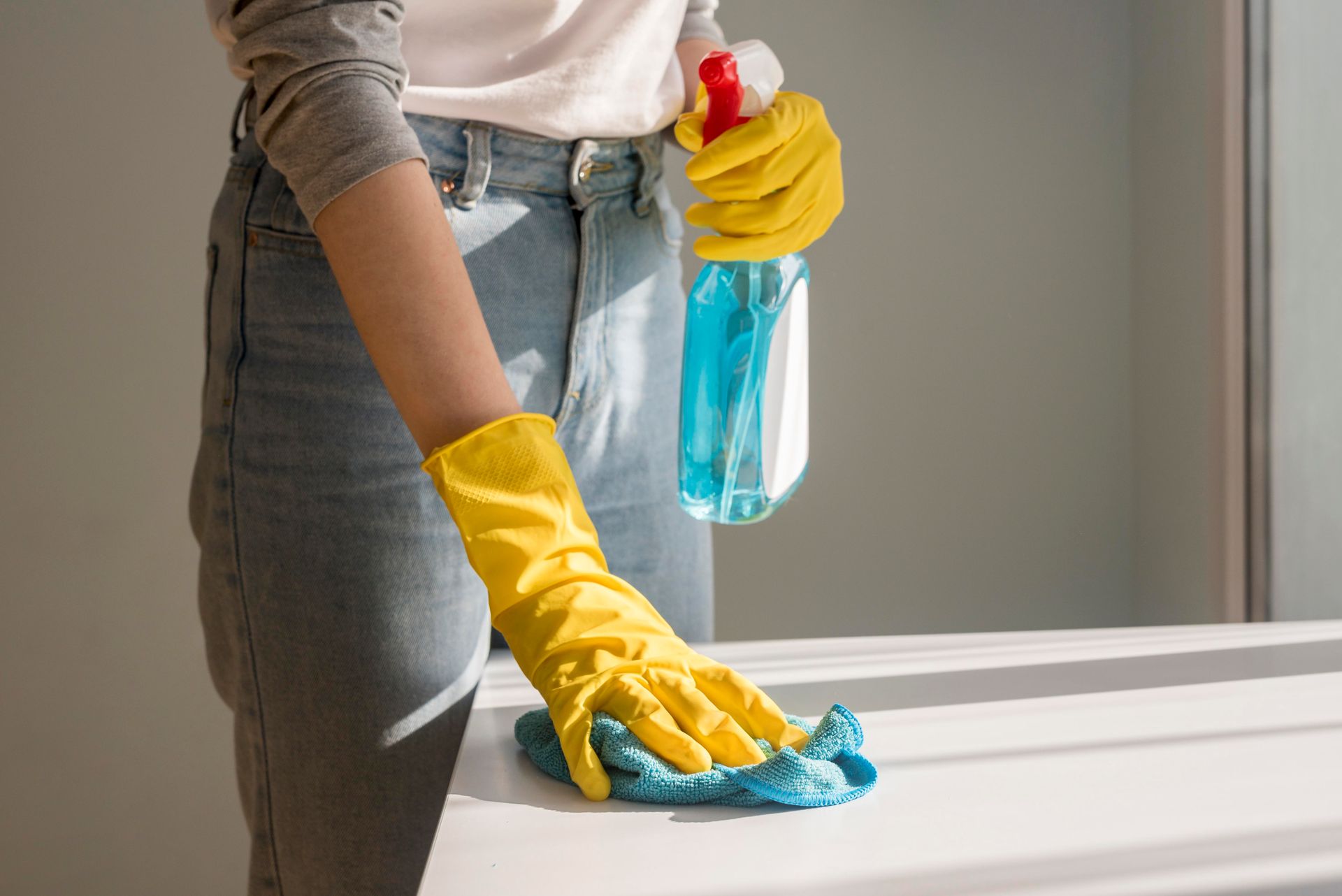 Woman in yellow gloves sprays and wipes a white surface with blue cloth.