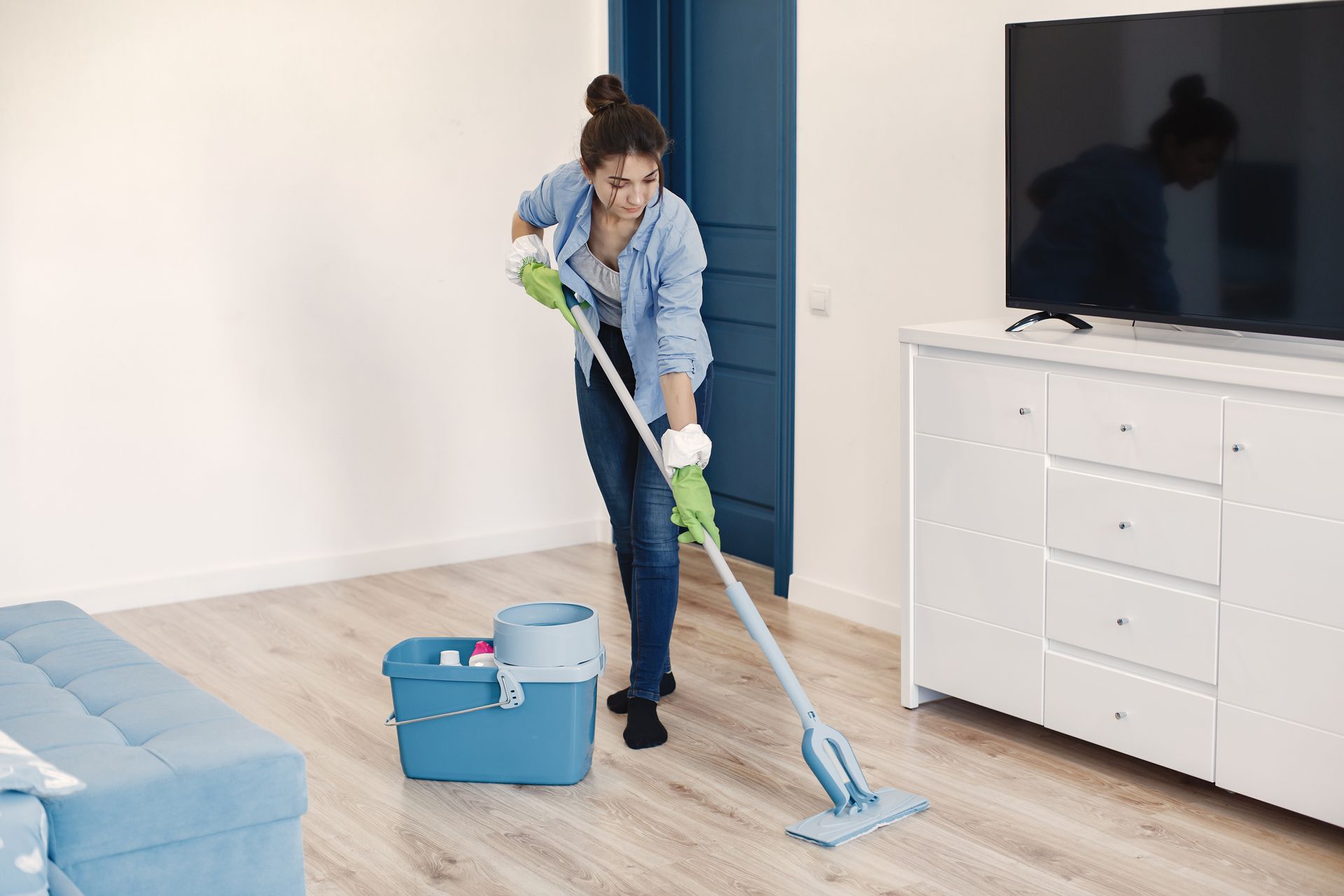 Woman mopping a wooden floor in a room with a blue bucket, TV, and white furniture.