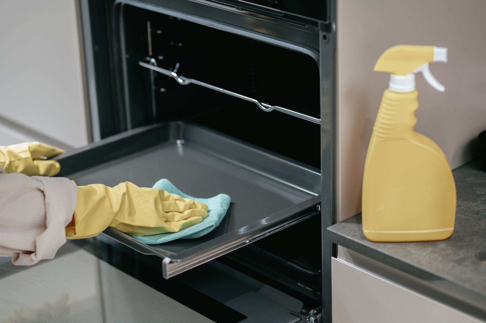 Person cleaning an oven with gloves, spray bottle on the counter.