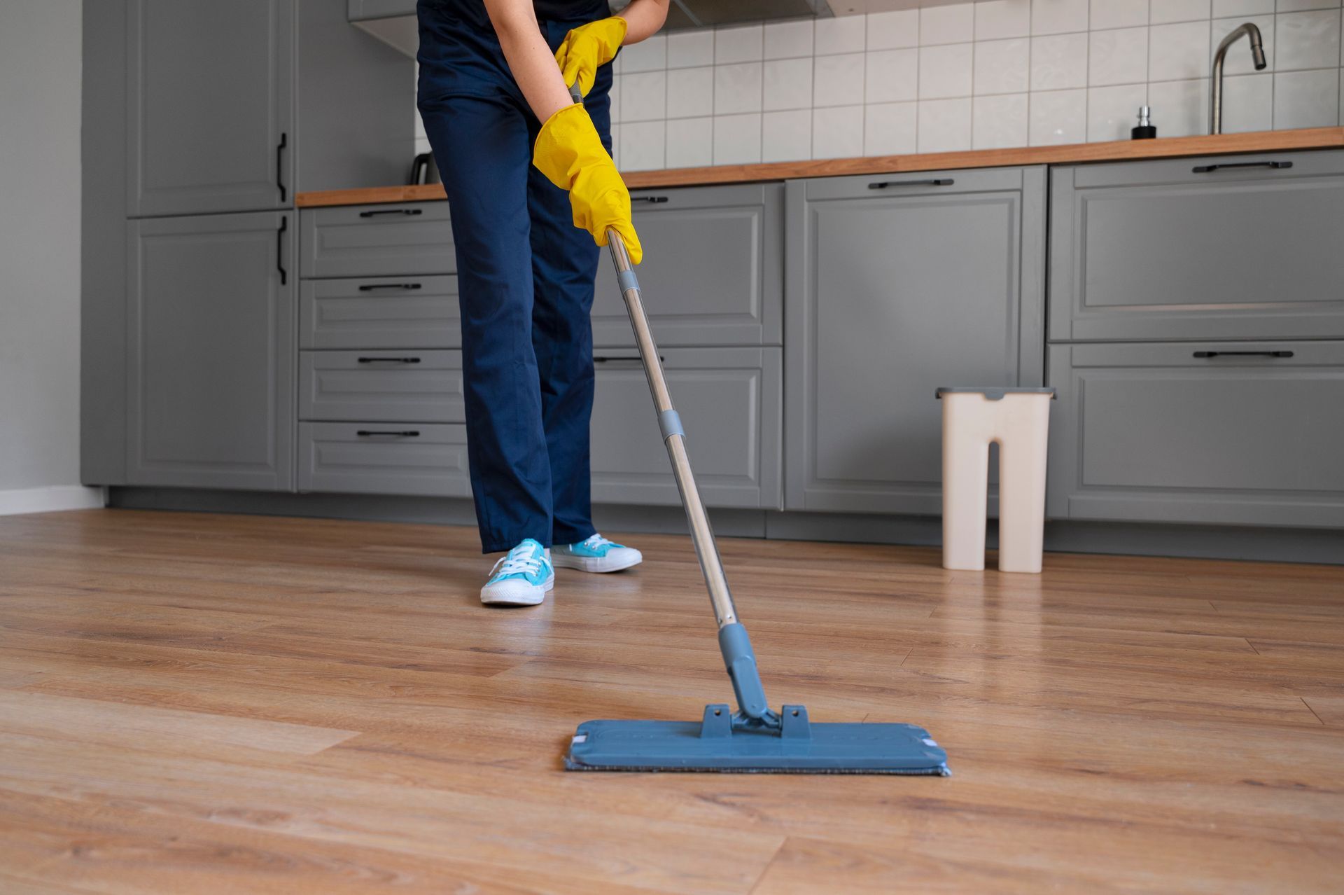 Person mopping a wooden floor in a kitchen; wearing blue jeans, yellow gloves, and blue sneakers.