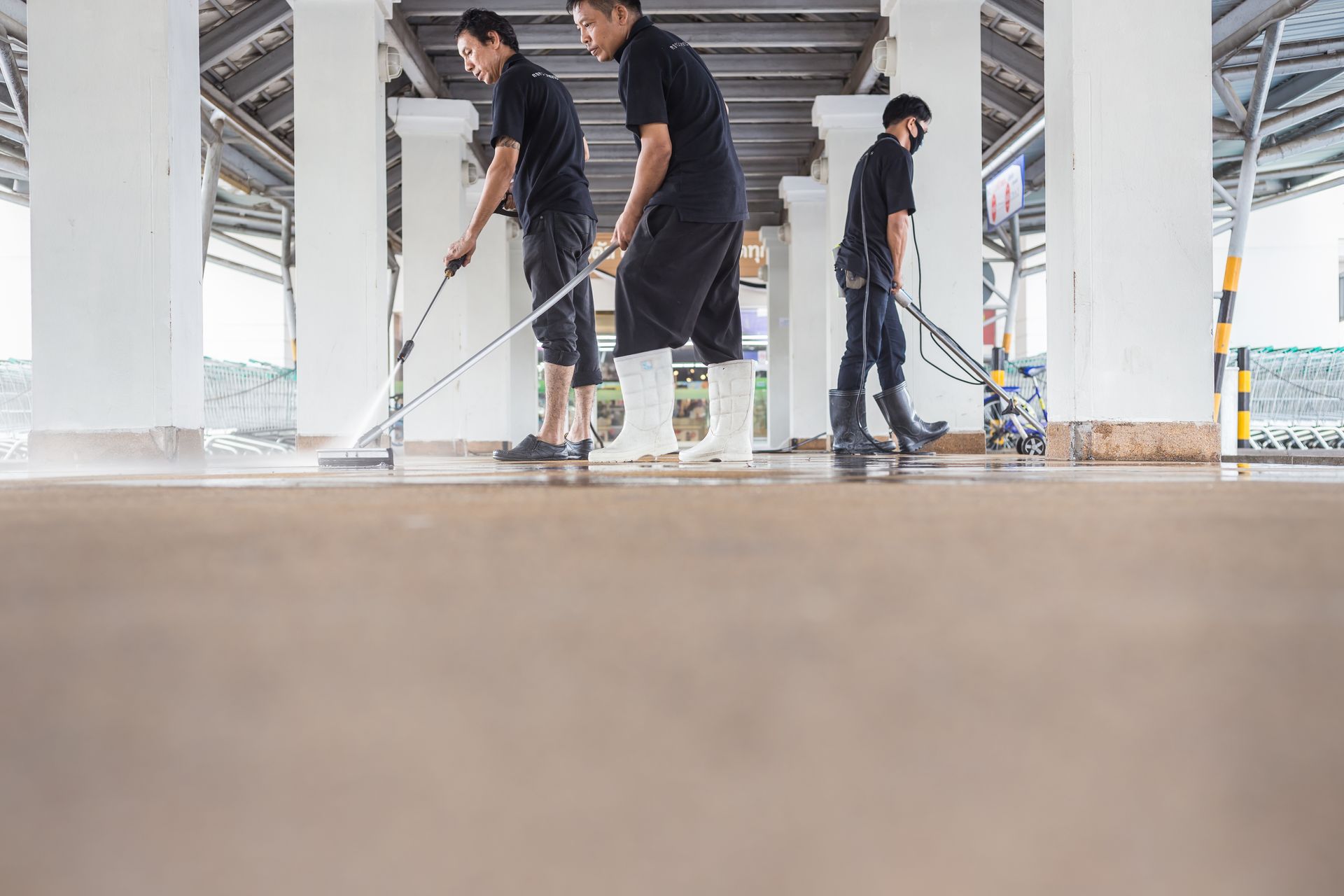 Three men in black uniforms cleaning a concrete floor outdoors with long-handled mops.