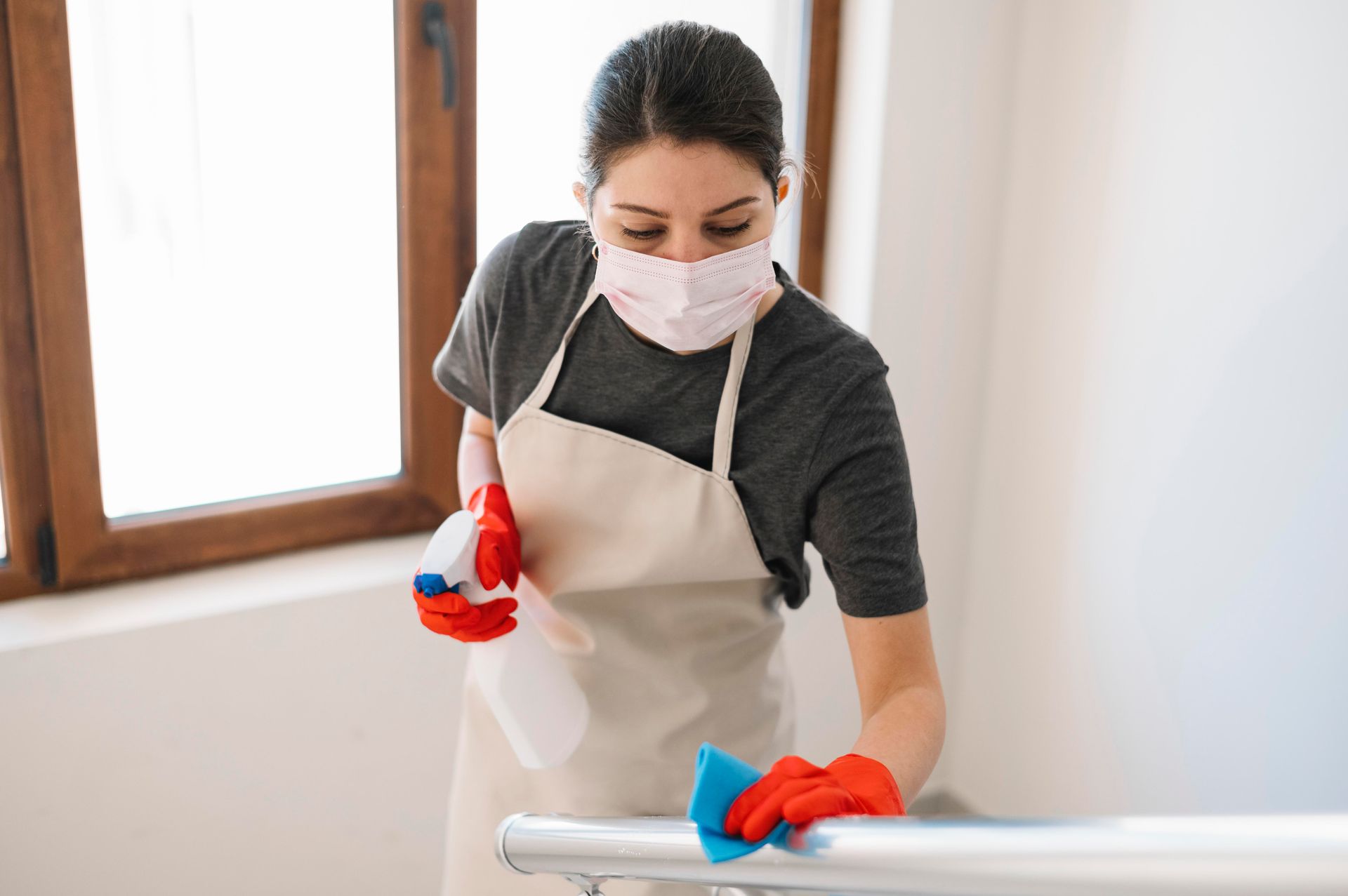 Woman wearing mask and gloves, cleaning a metal handrail with a spray bottle and sponge.