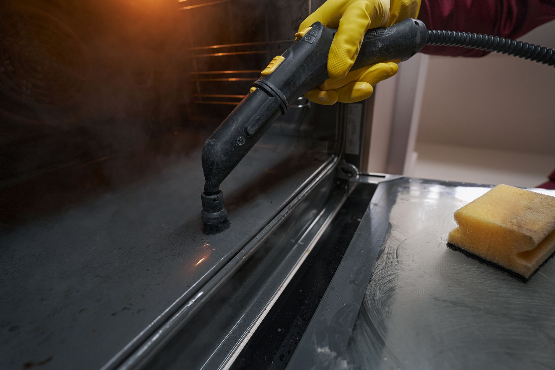 Person wearing yellow gloves steam-cleaning a shiny metal surface inside an oven.