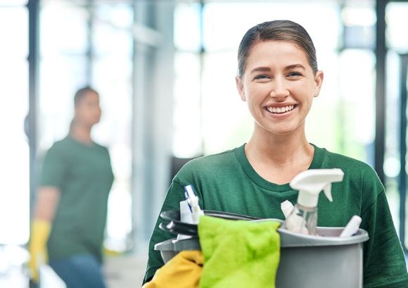 A woman is holding a bucket of cleaning supplies and smiling.