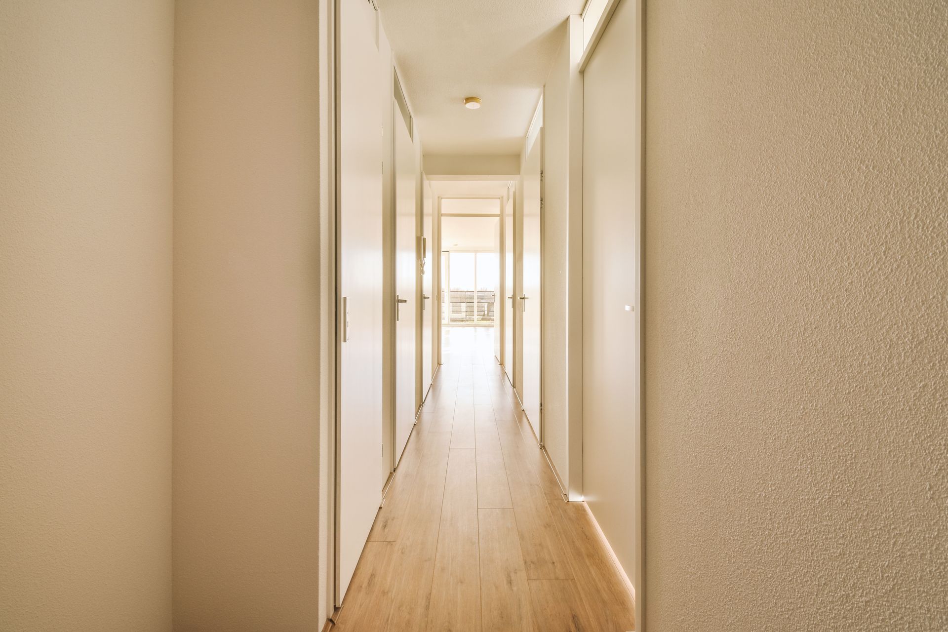 Long hallway with closed white doors and light wooden floor leading to a bright window.