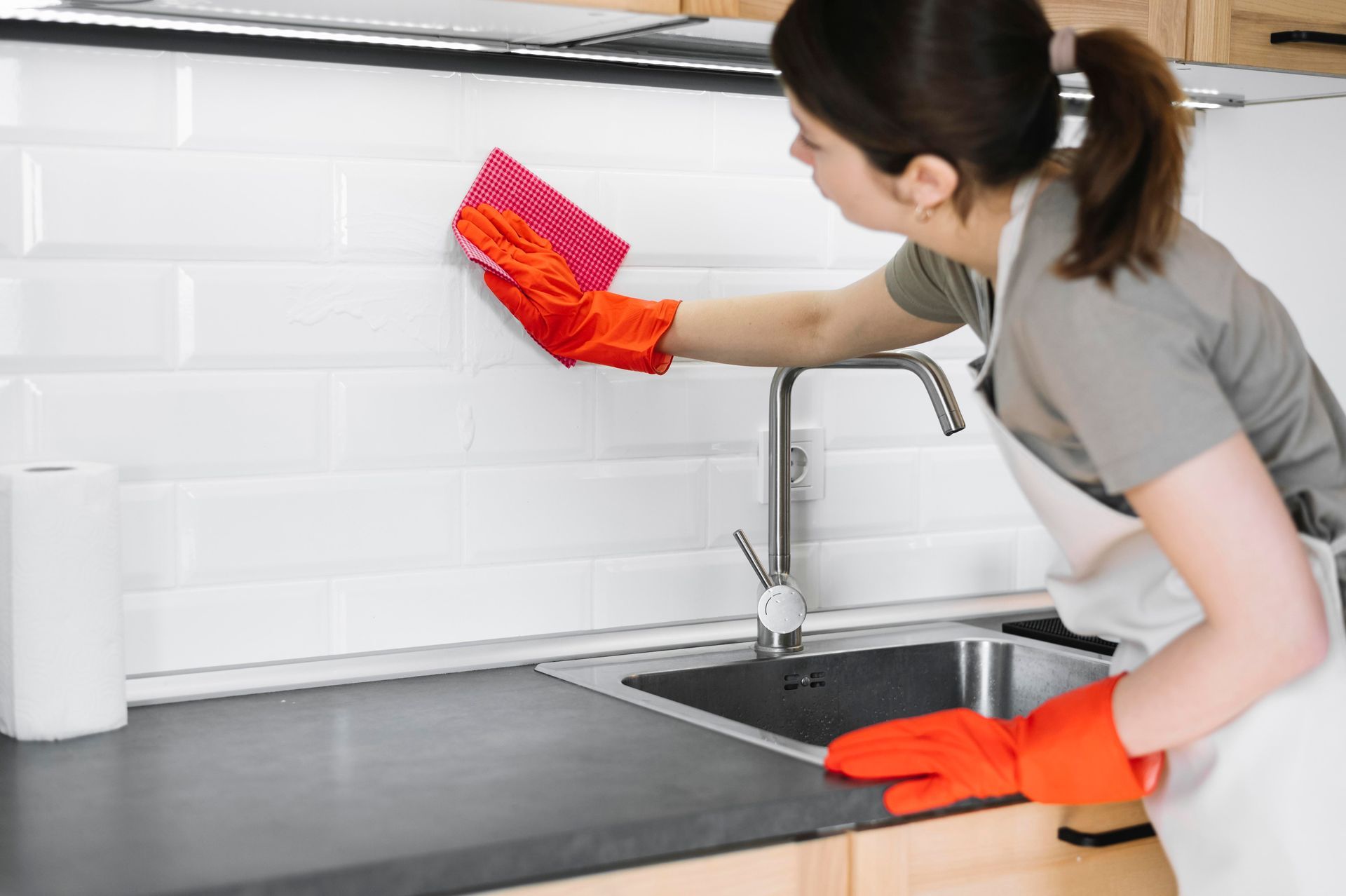 Woman wearing orange gloves and an apron cleaning white tiled backsplash above a kitchen sink.