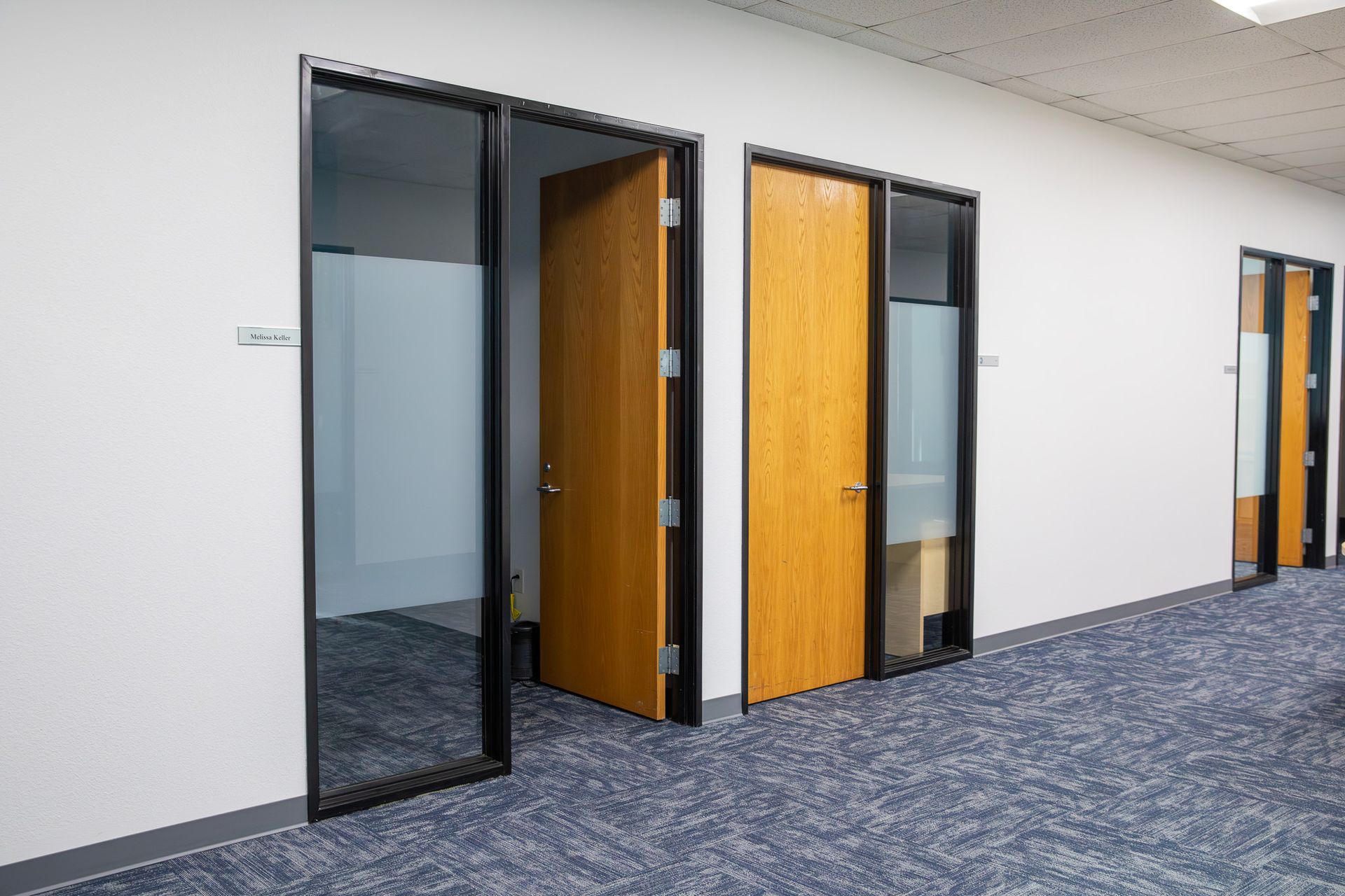 Hallway with three open doors, two with wooden doors and one with glass. Dark frames. Blue carpet. White walls.