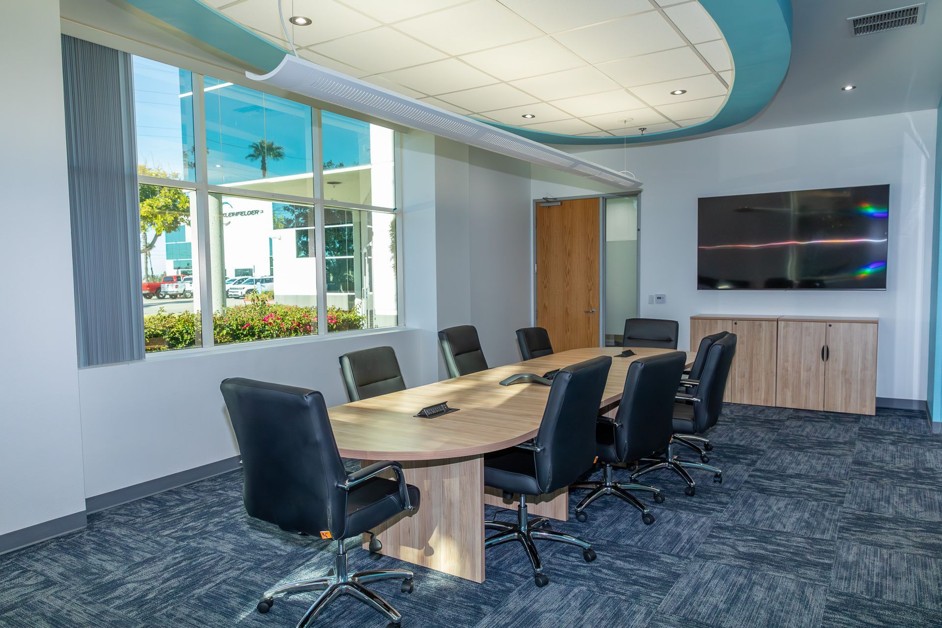 Meeting room with oval table, black chairs, large window, TV, and blue and white accents.