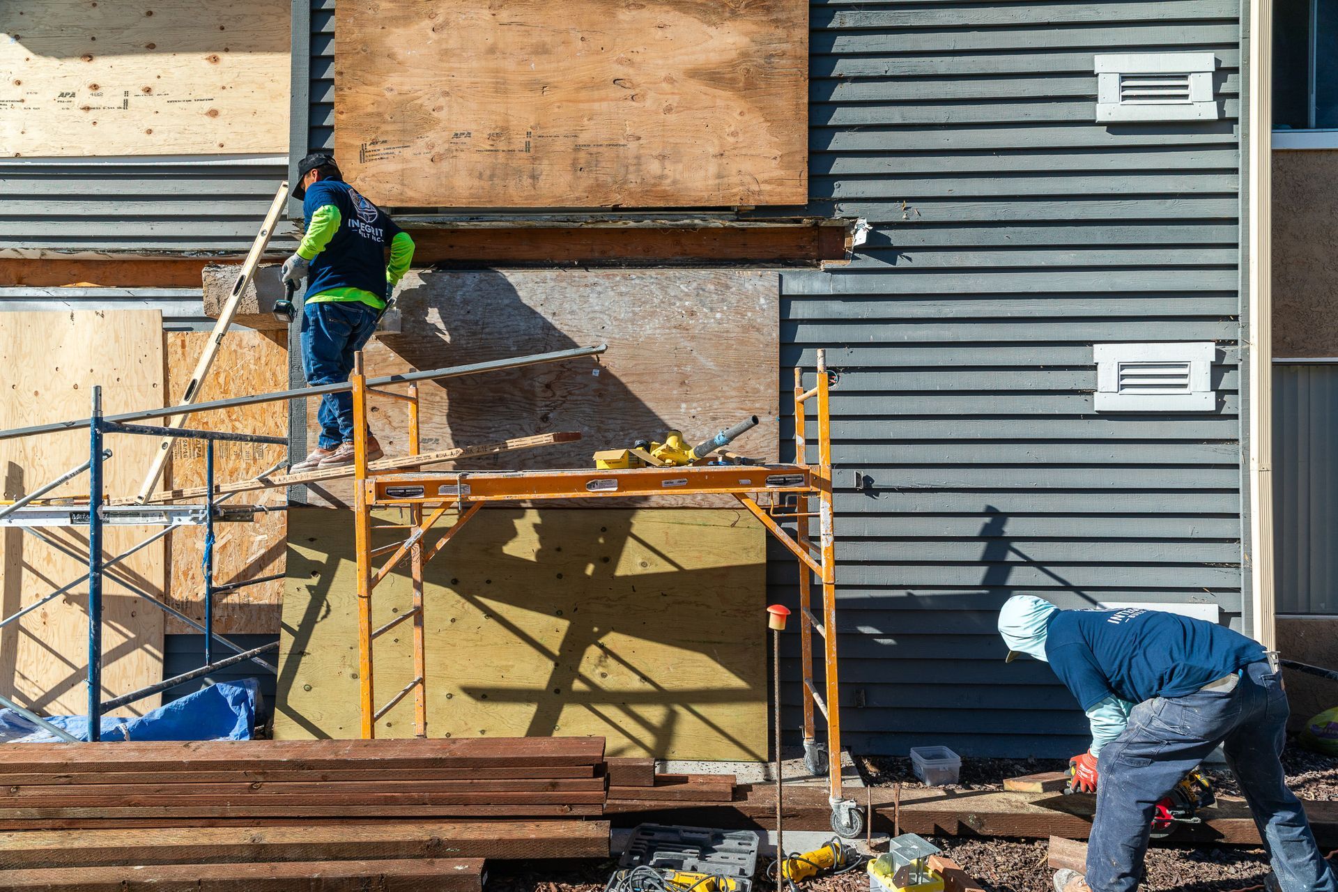 Construction workers repairing a building. One on scaffolding, one on the ground, plywood and tools visible.