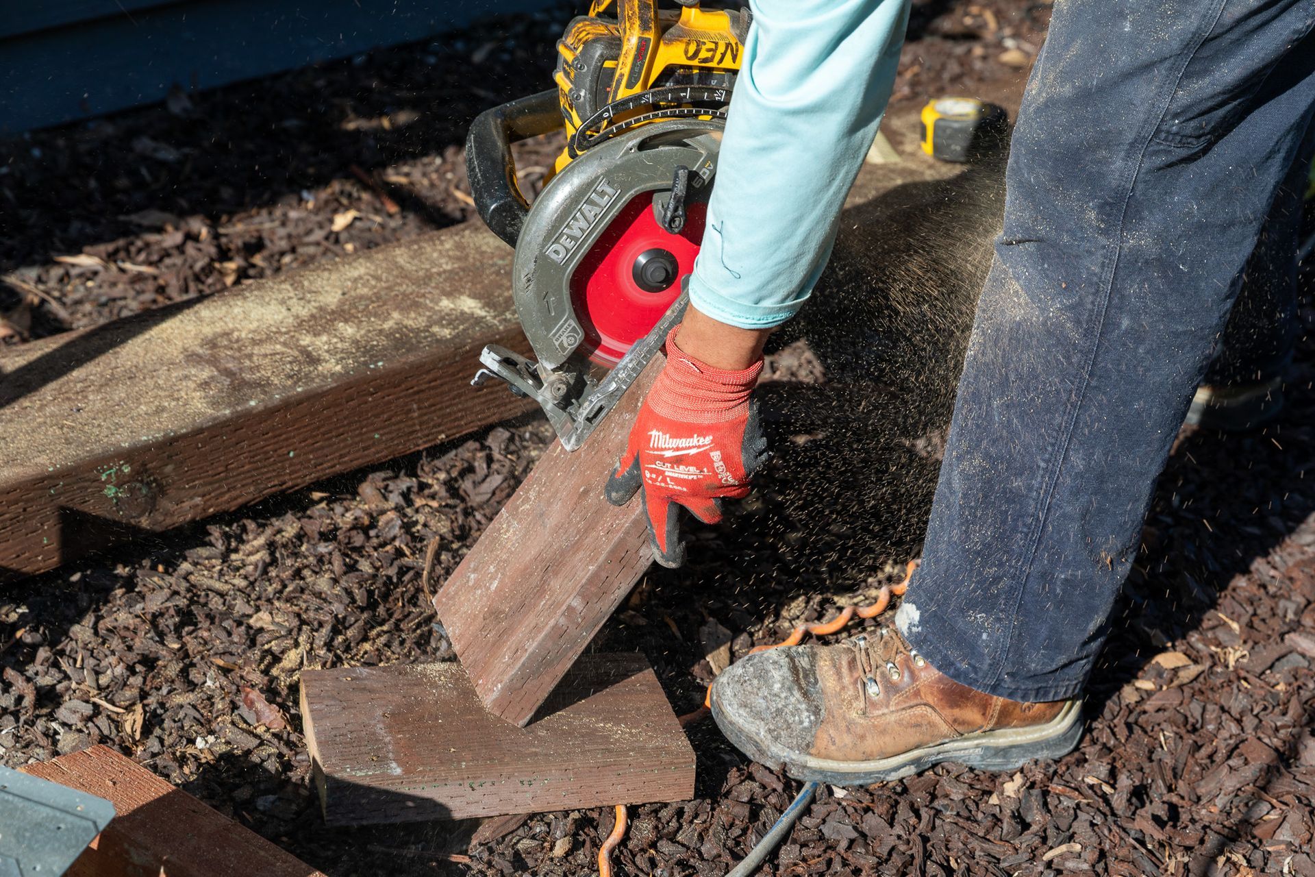 Person using a circular saw to cut a wooden plank outdoors; sawdust visible.