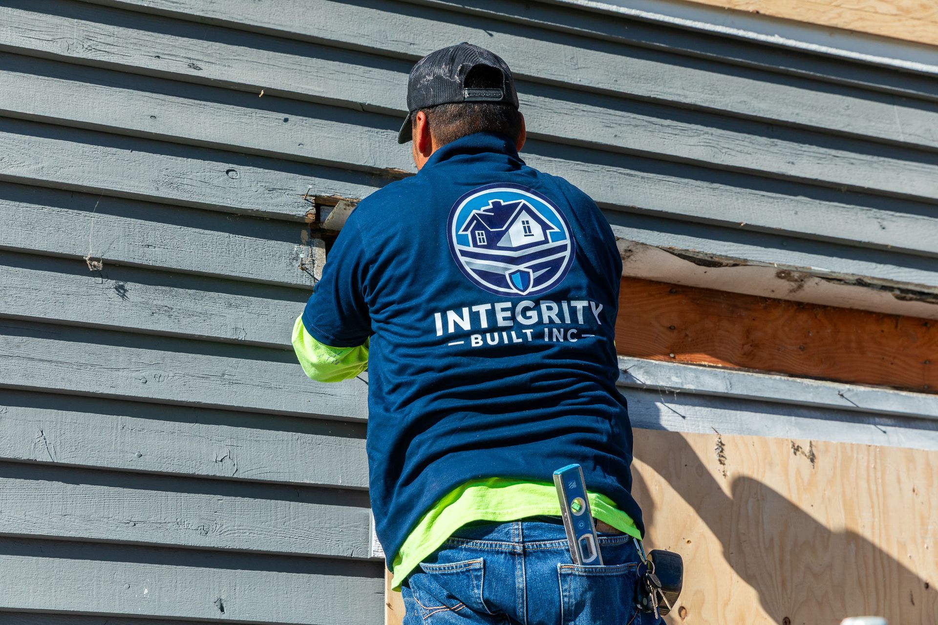 Construction worker removing siding, wearing blue shirt with company logo, on a building exterior.