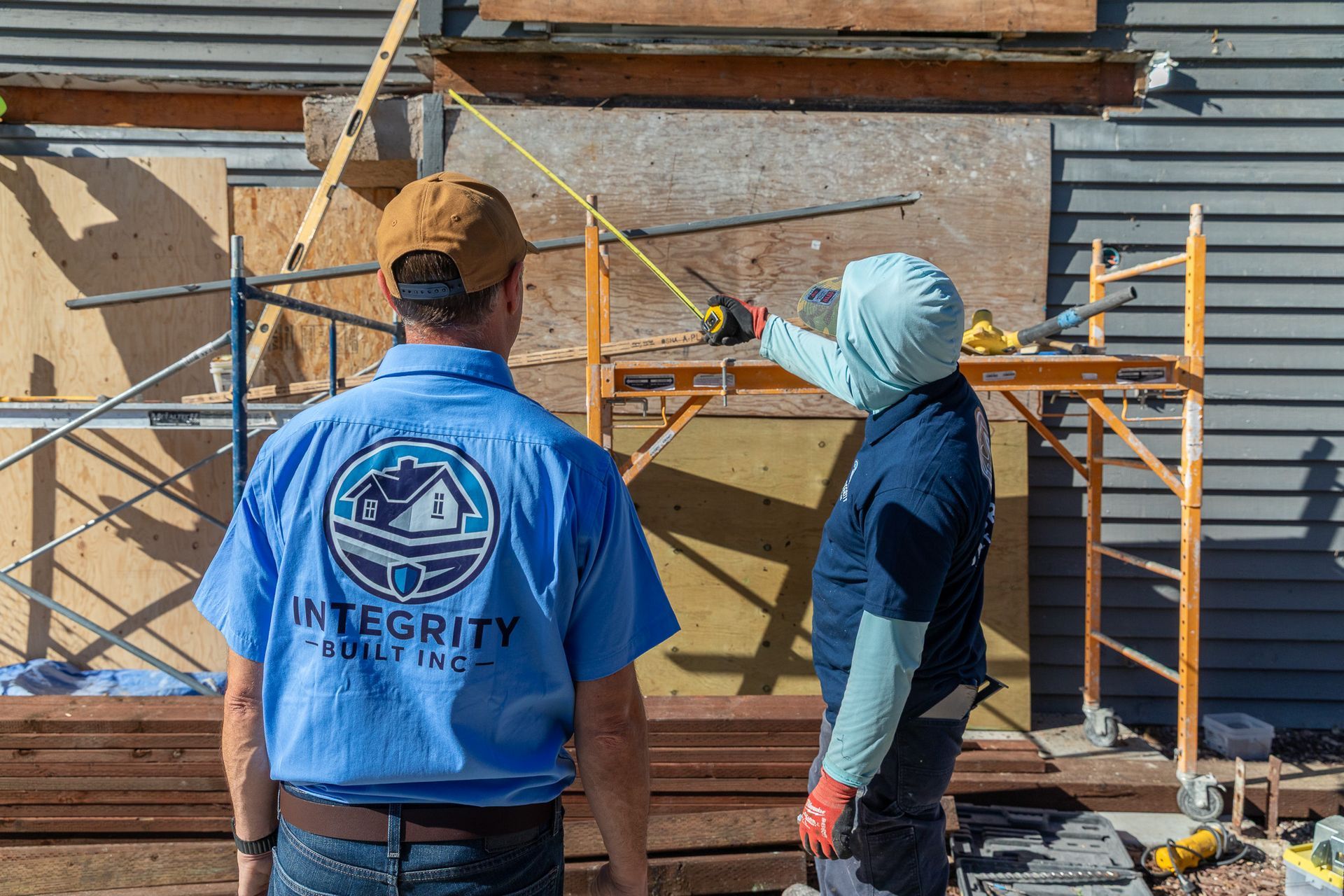 Two construction workers measure a boarded-up window opening. One points a tape measure. Blue shirts, exterior setting.