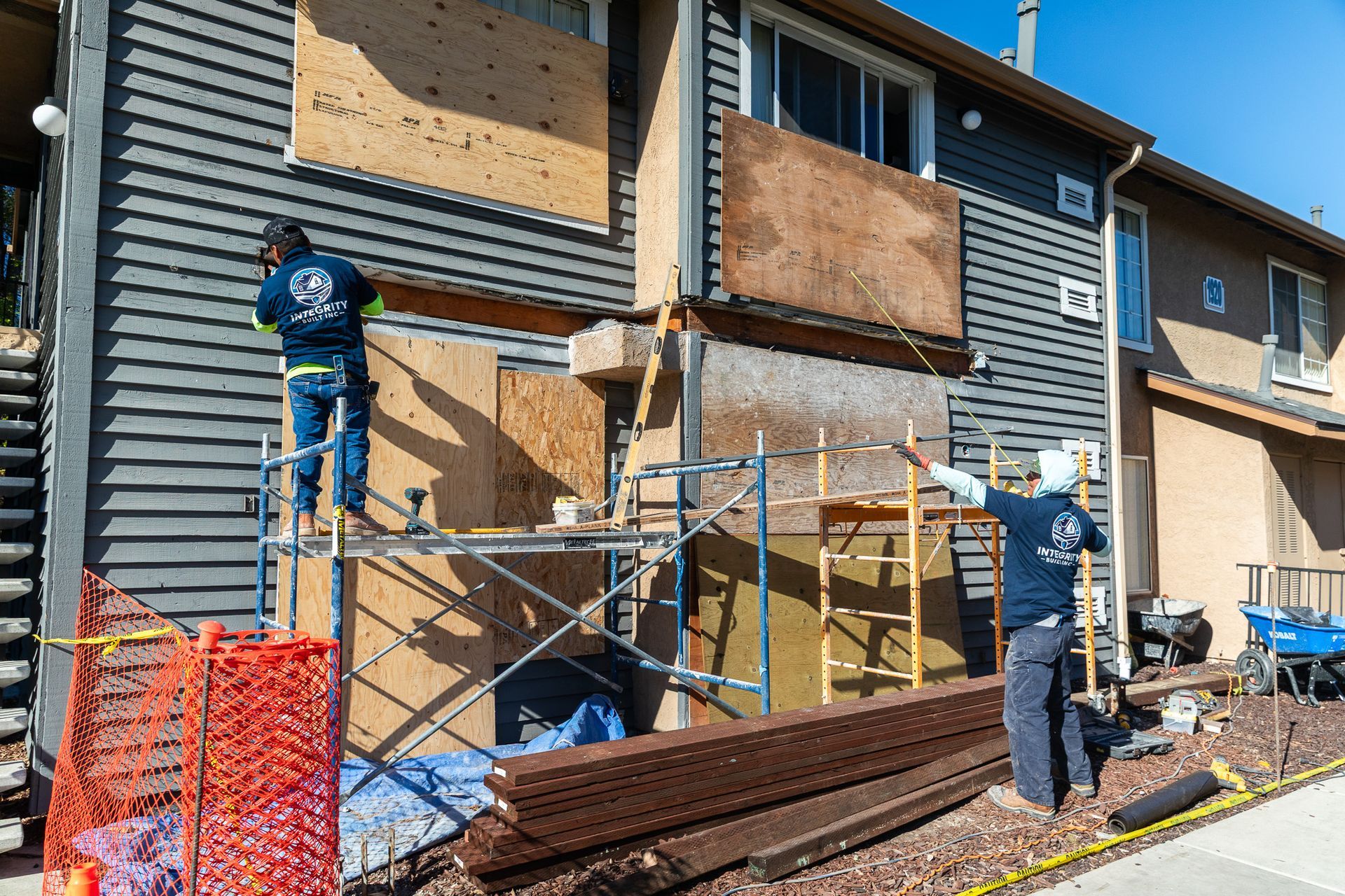 Workers boarding up a building with plywood, scaffolding in place. Orange barrier on the left.
