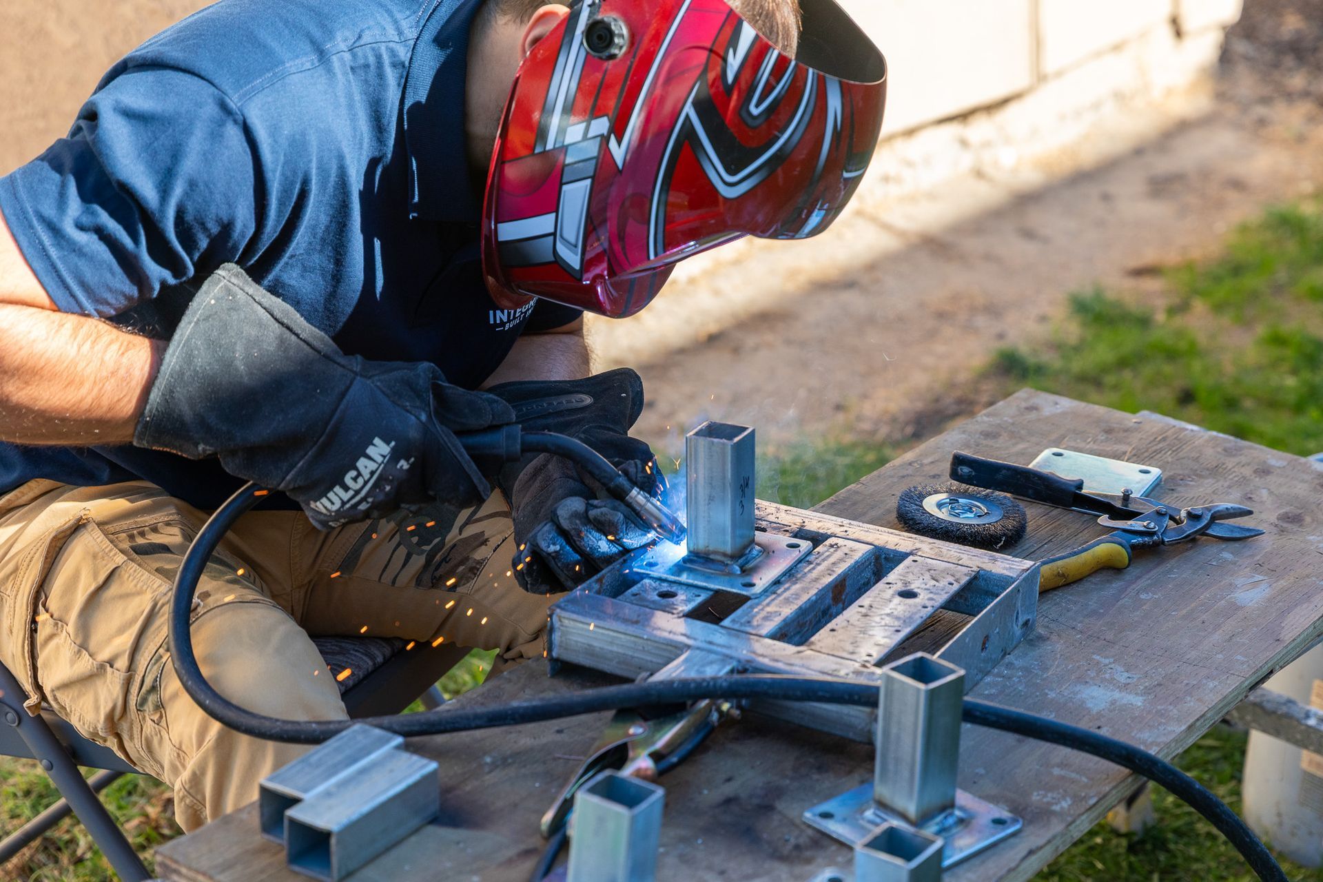 Person welding metal pieces outdoors, wearing a red welding helmet and gloves.