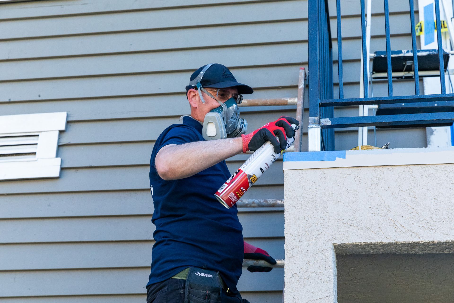 Man in respirator sprays paint on a concrete ledge next to blue siding and a railing.