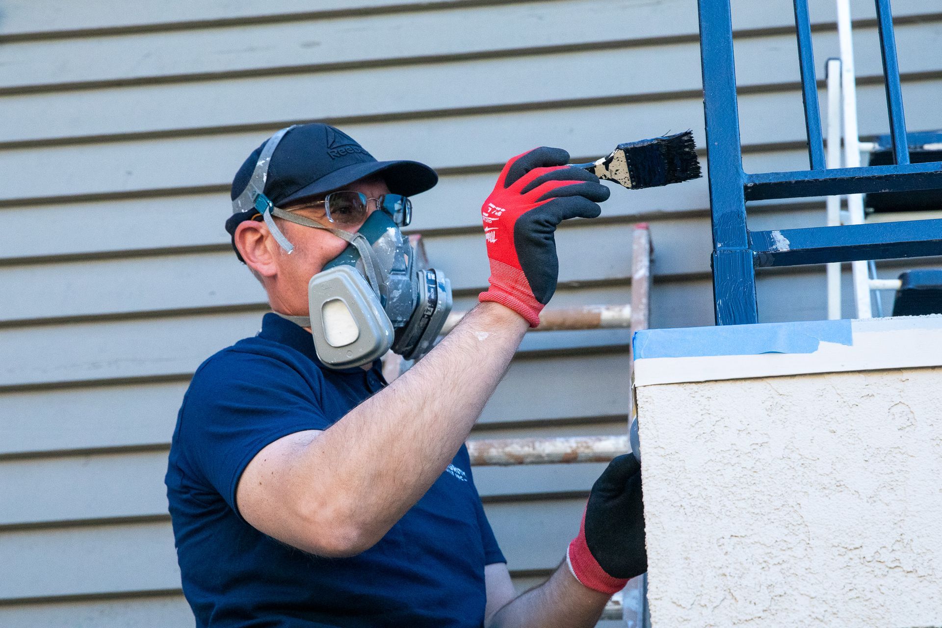 Man wearing a respirator and gloves paints a blue railing on a white surface, outdoors.