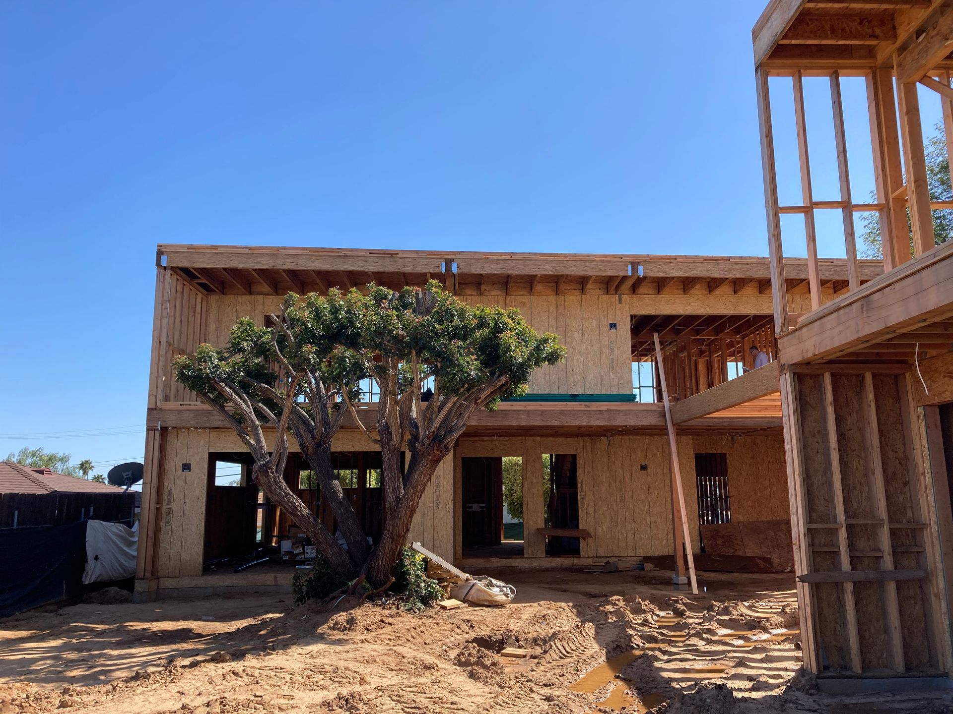 Construction of a two-story wooden house on a dirt lot, with a mature tree in front under a blue sky.