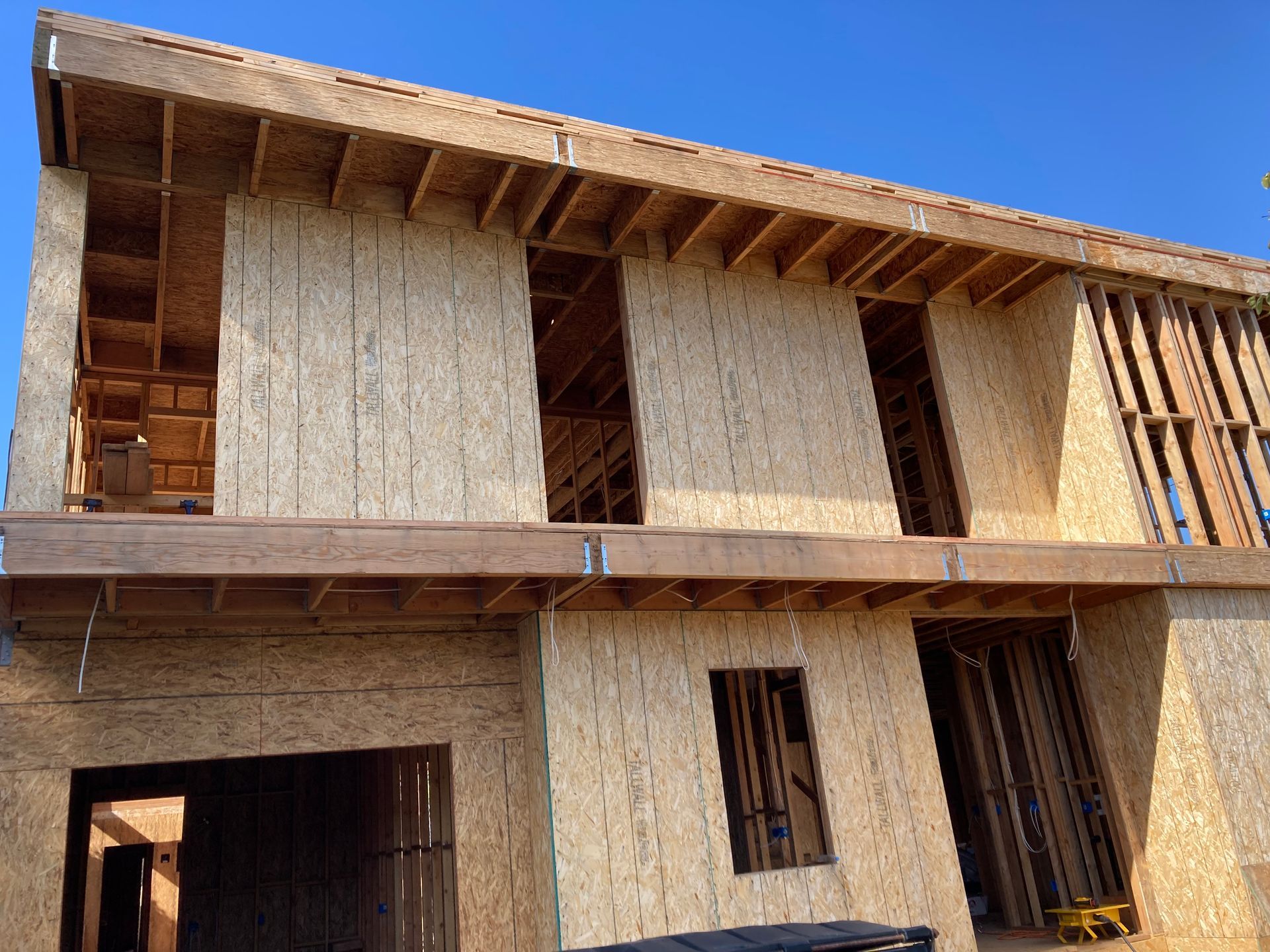 Two-story house under construction with exposed wooden frame and OSB sheathing against a blue sky.