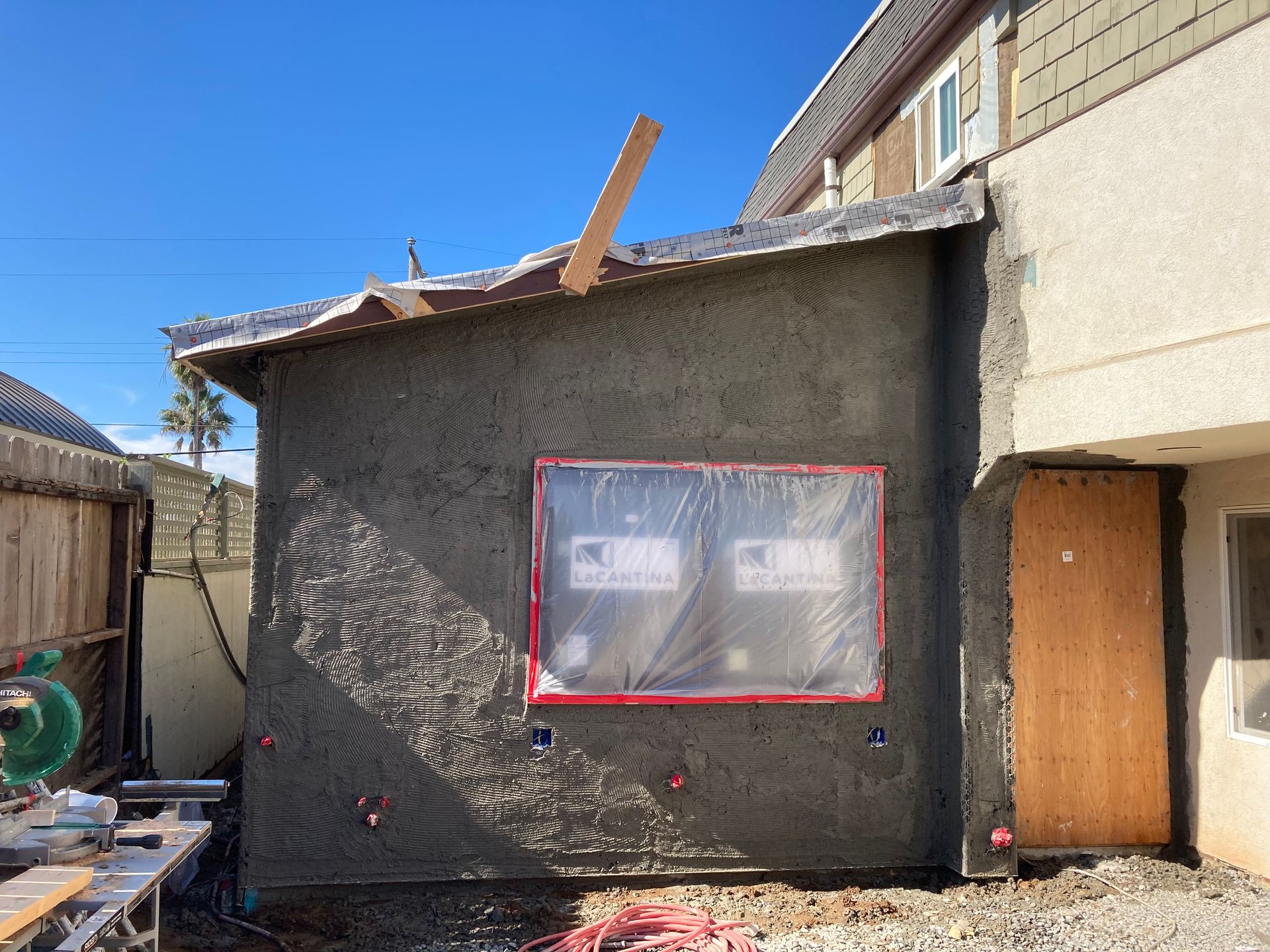 Exterior construction, stucco finish, window covered in plastic, wooden door, sunny day.