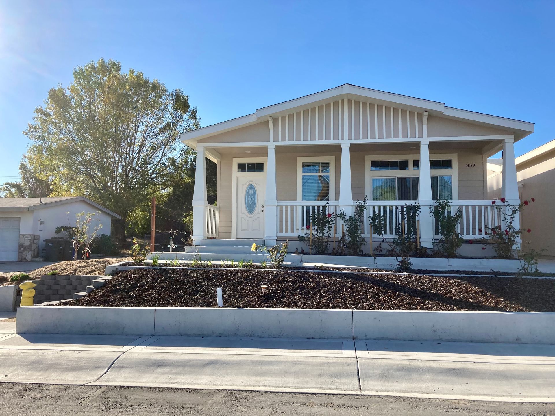 Beige bungalow with a white porch and railing, set on a raised bed with mulch, under a blue sky.