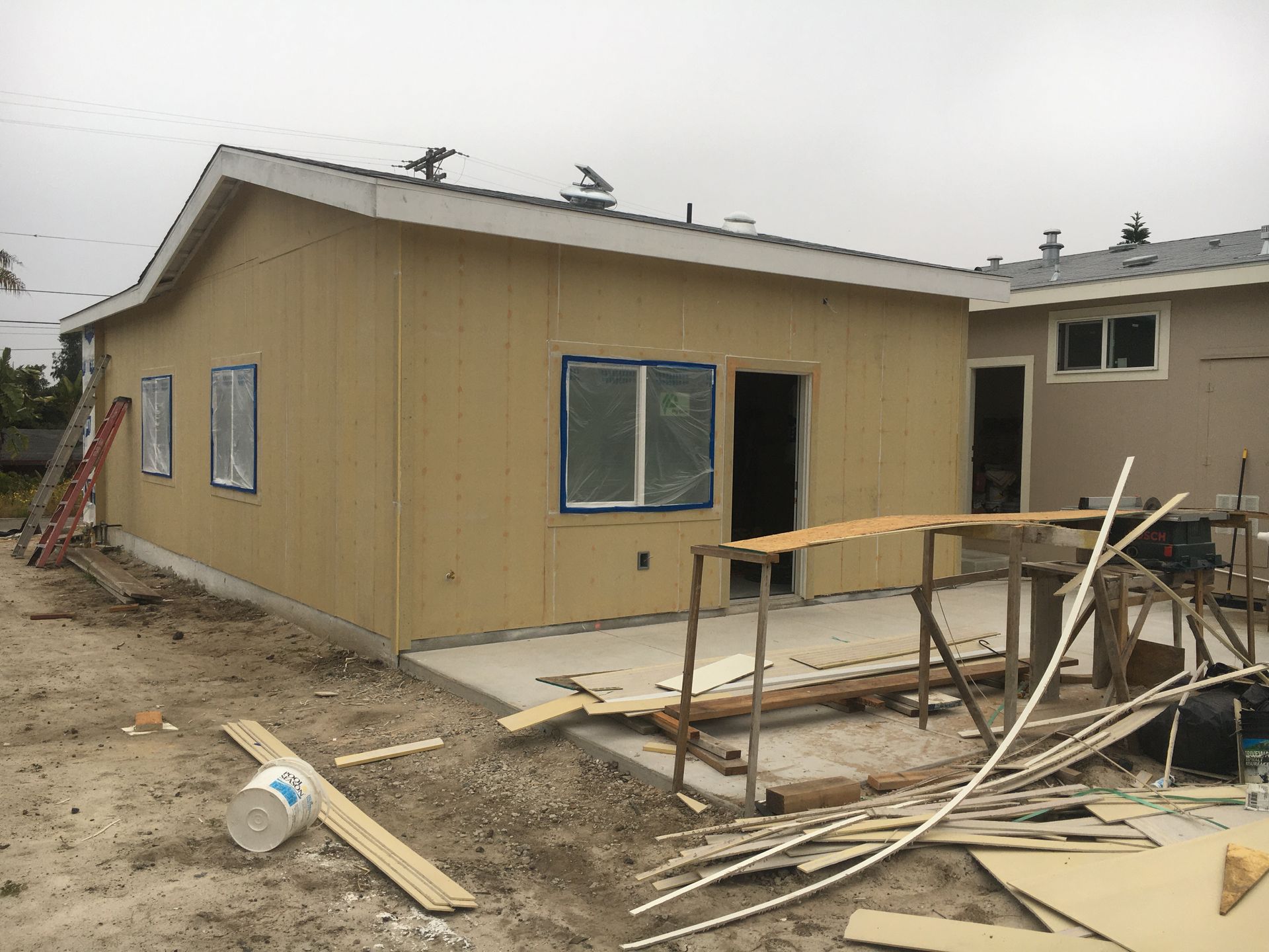 Exterior of a home under construction; beige siding, windows, open doorway, concrete patio, construction materials.