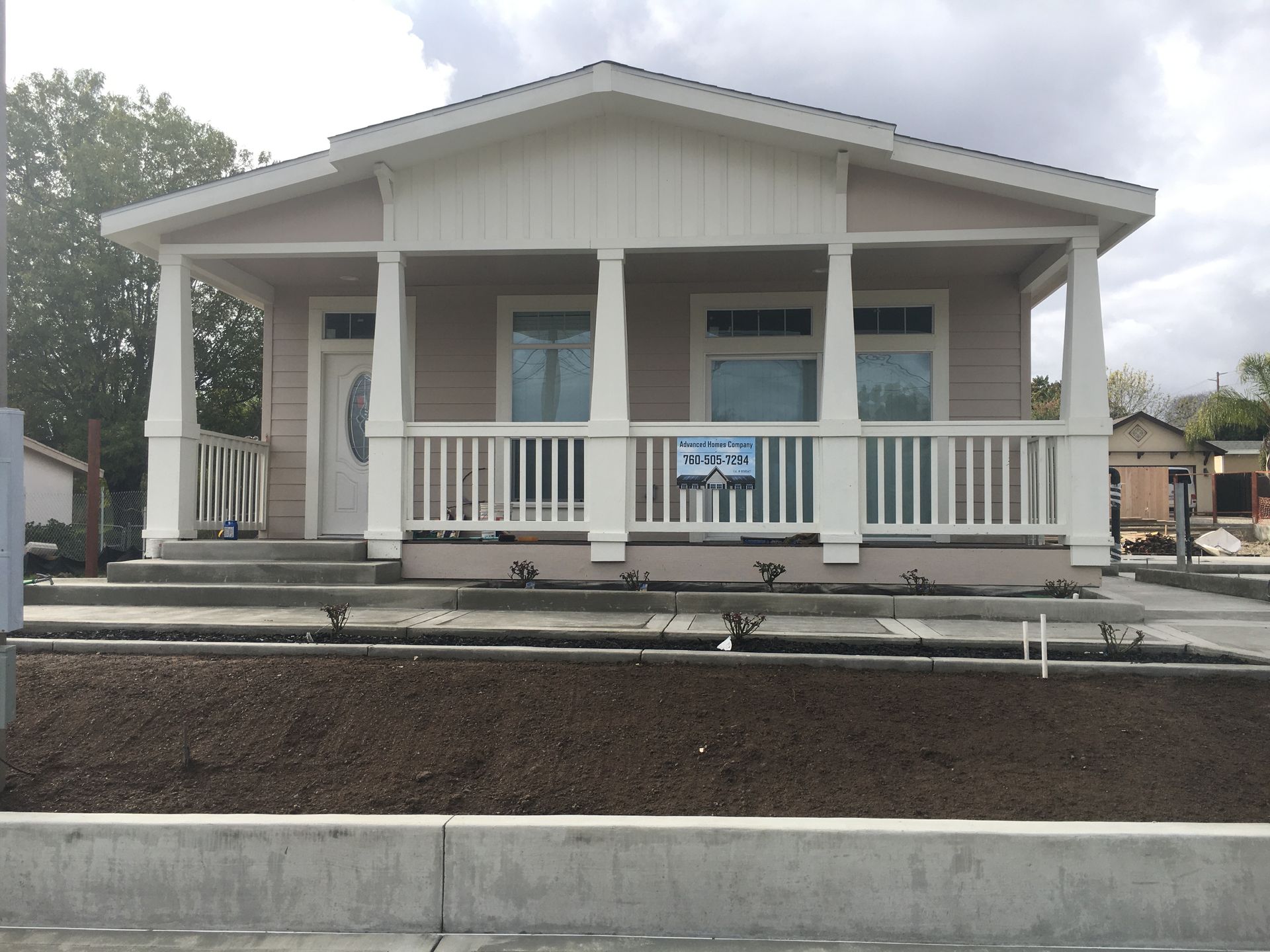 Light pink house with white trim, porch, and railing, set in a dirt landscape next to a sidewalk and a curb.