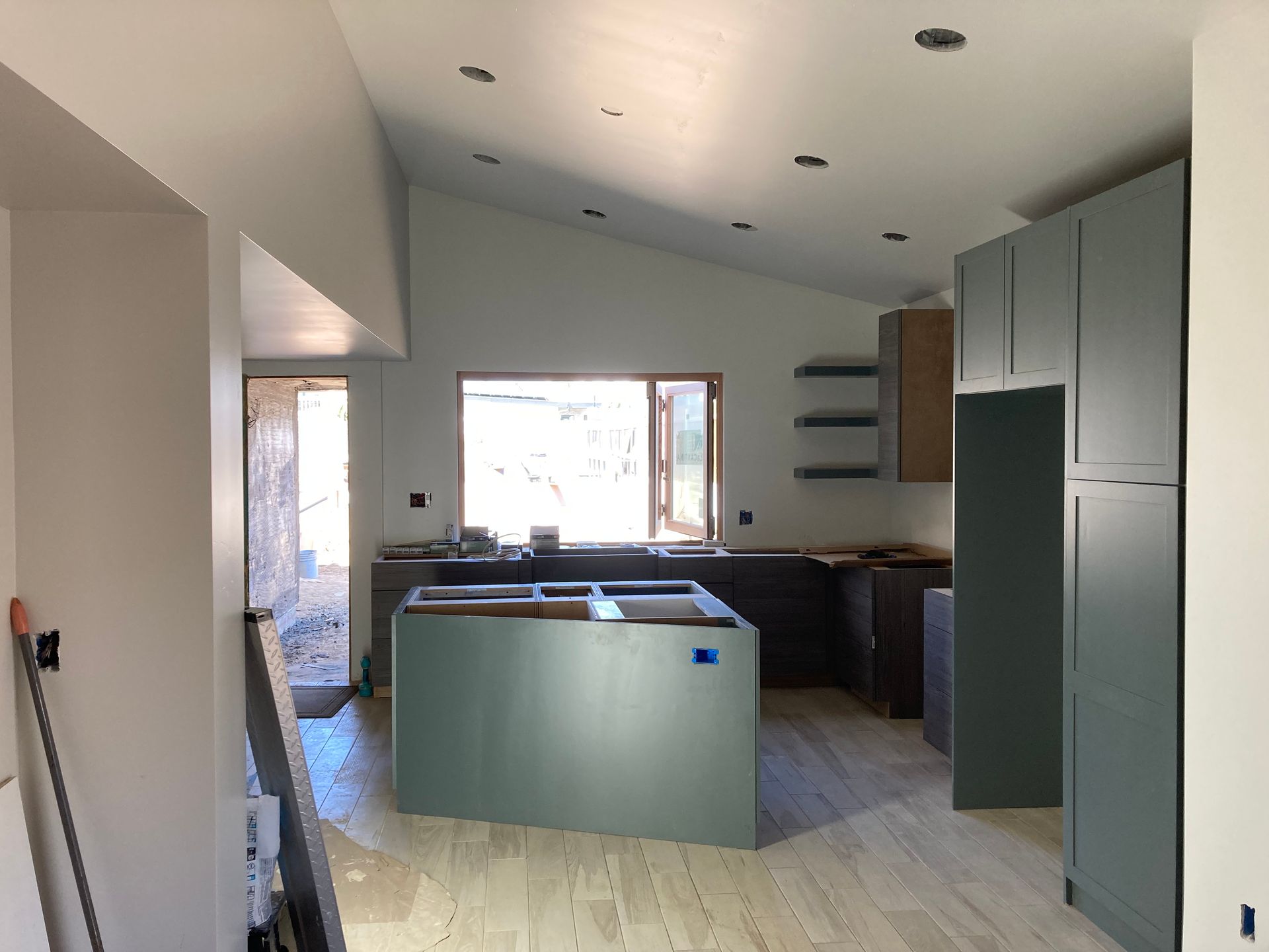 Kitchen under construction with light wood floors, blue-green cabinets, and a central island.