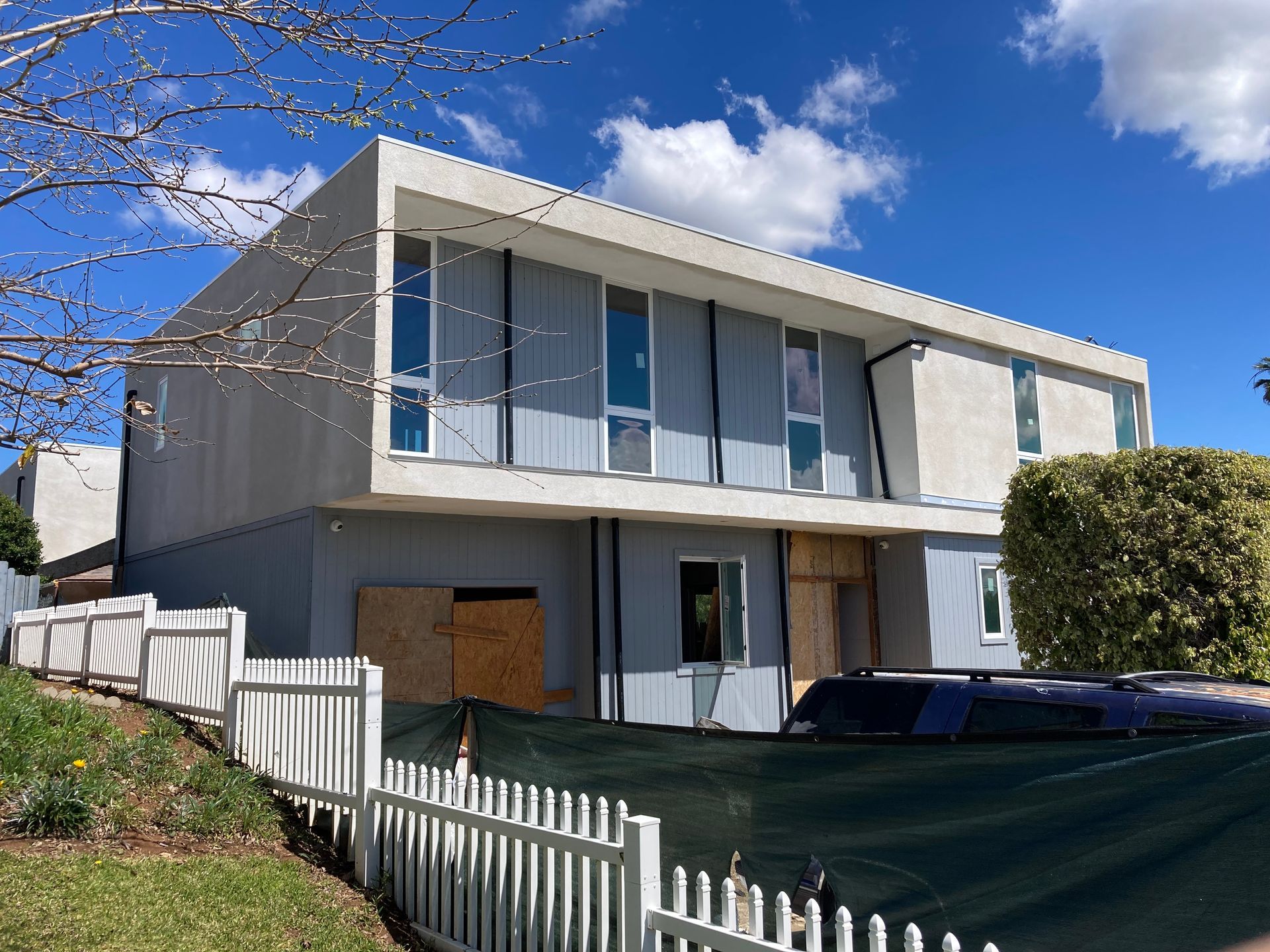 Two-story house undergoing renovation; light blue siding, boarded windows, white fence in front.