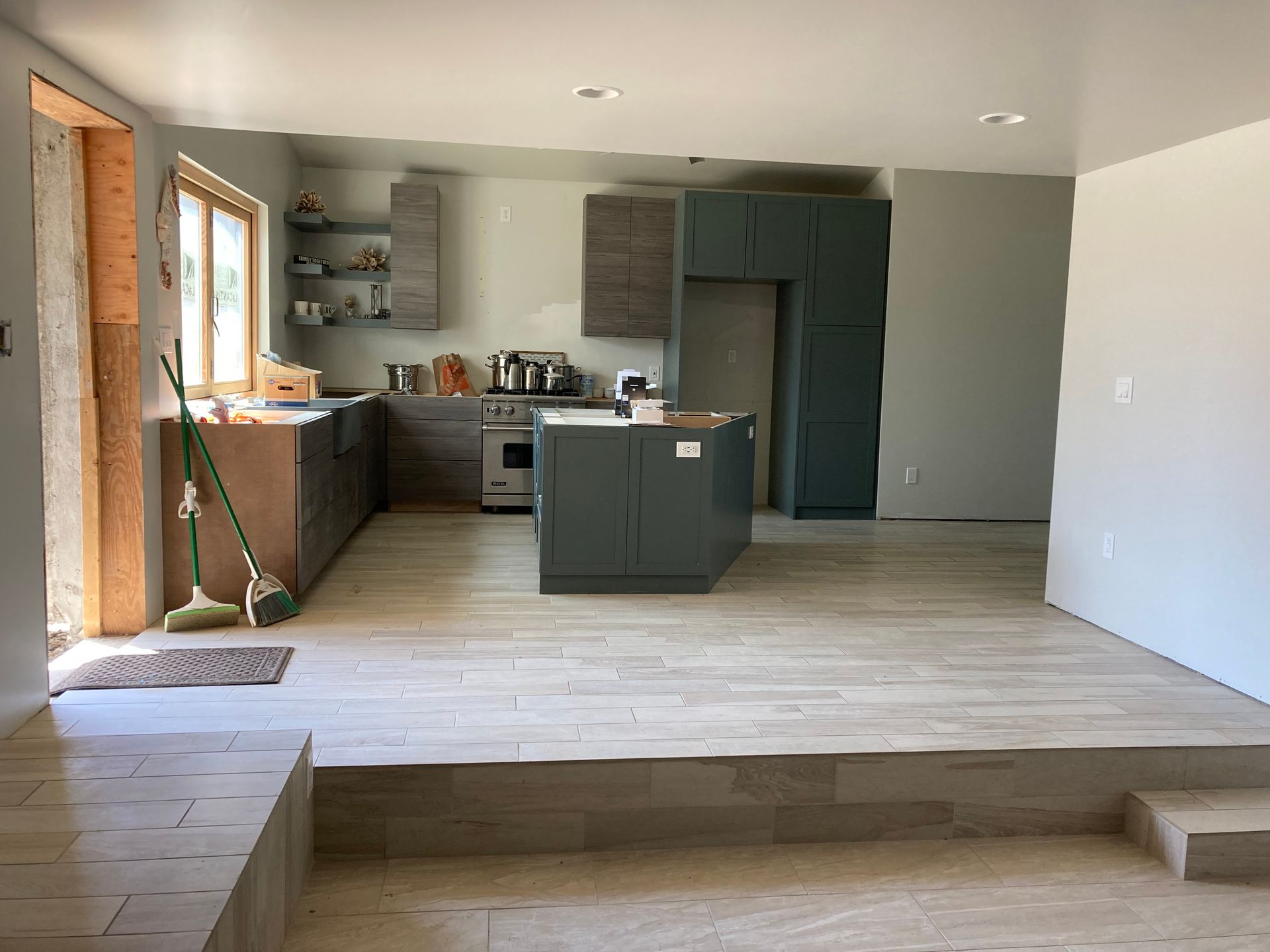 Kitchen renovation with two-tiered flooring. Gray cabinets and island, unfinished walls, and natural light.