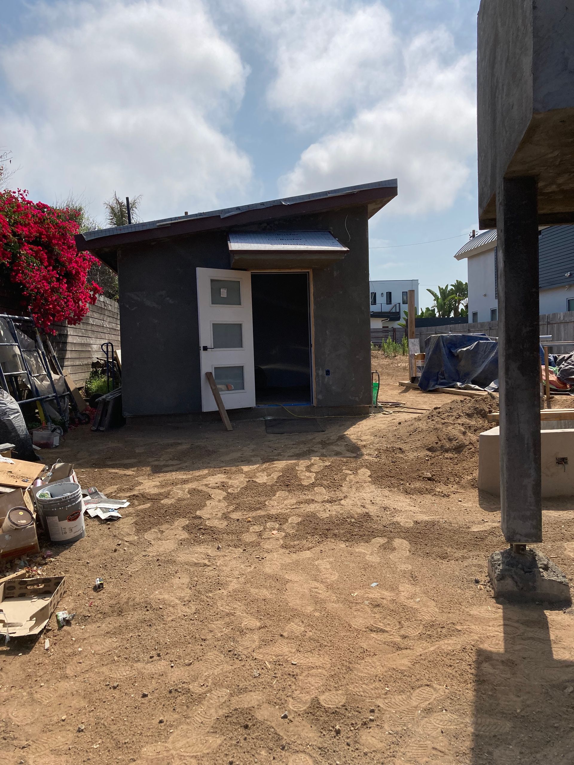 Small stucco building with a slanted roof and open door on a dirt lot; bright sky in background.