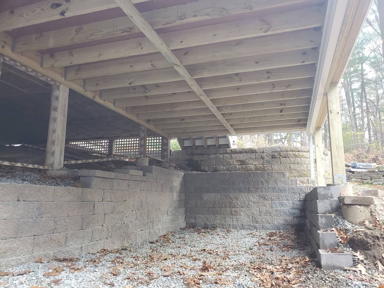 Underside of a wooden deck supported by concrete block walls and pillars. Gravel ground below, steps on the right.