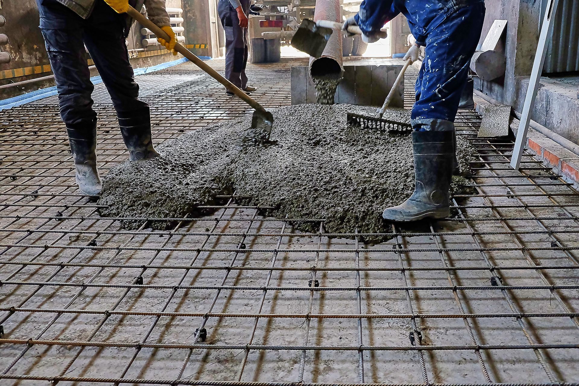Two construction workers are pouring concrete on a steel mesh floor.