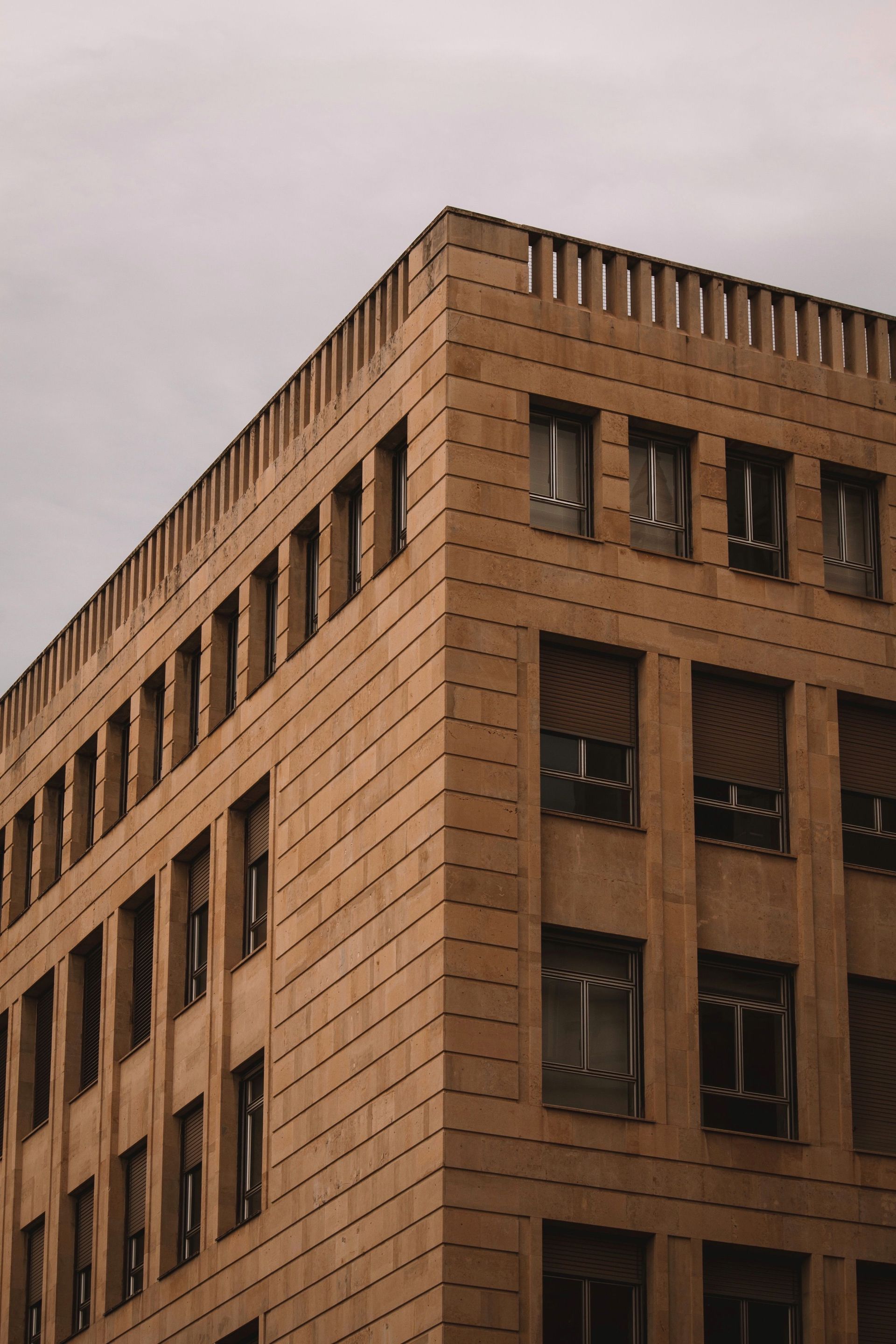 Tan brick office building corner with rows of dark windows under an overcast sky