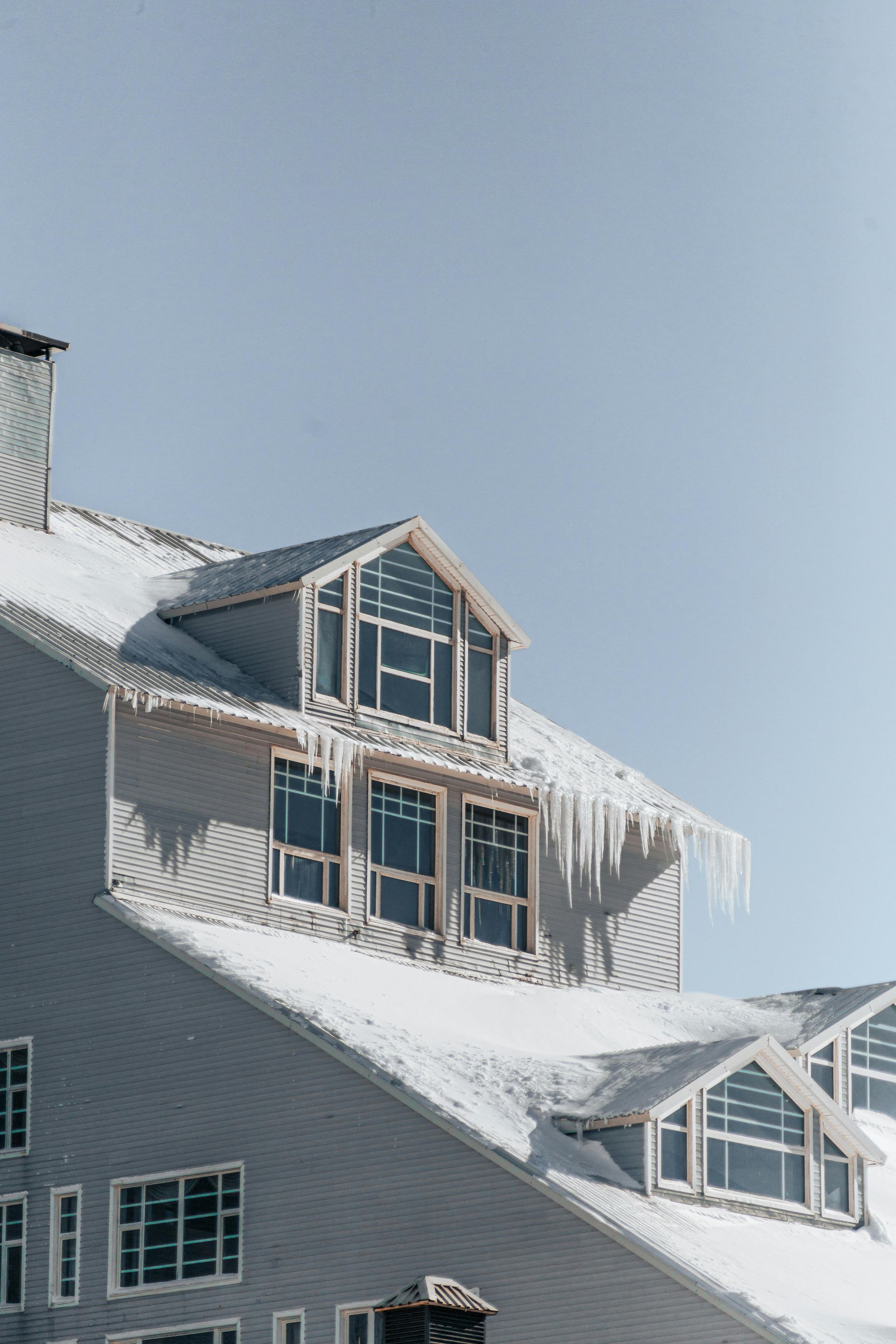 A gray building with a snowy roof, multiple dormer windows, and large icicles hanging from the eaves under a clear sky.