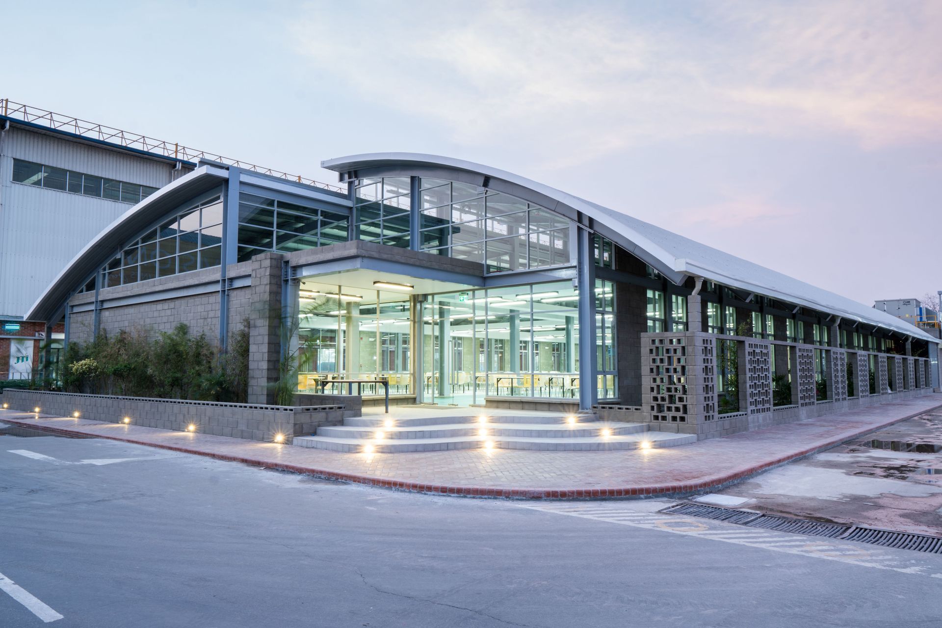 Modern glass-fronted building with curved roof and lit entrance at dusk
