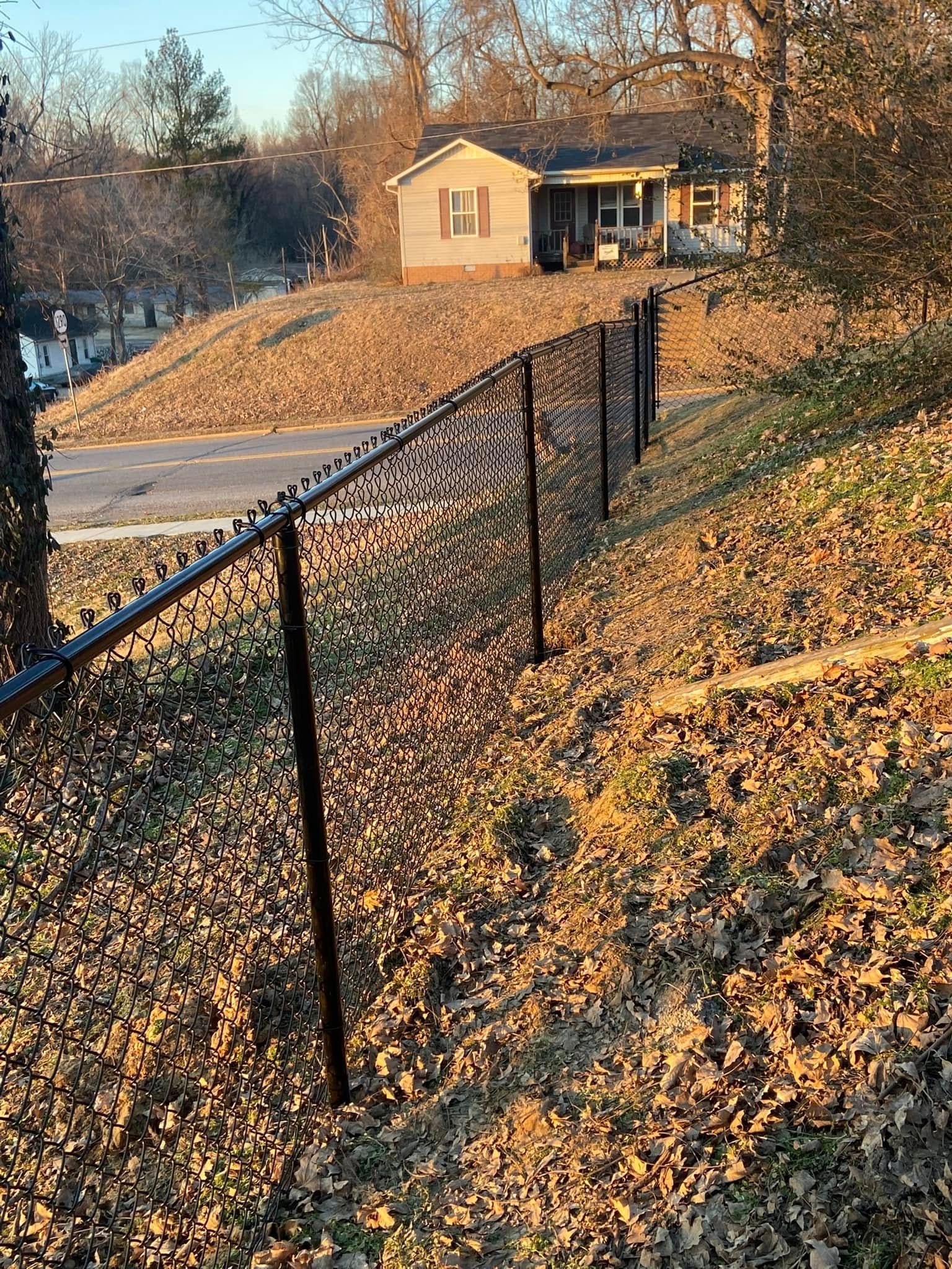 Black chain-link fence on a hillside, leading to a small house. Autumn leaves cover the ground.