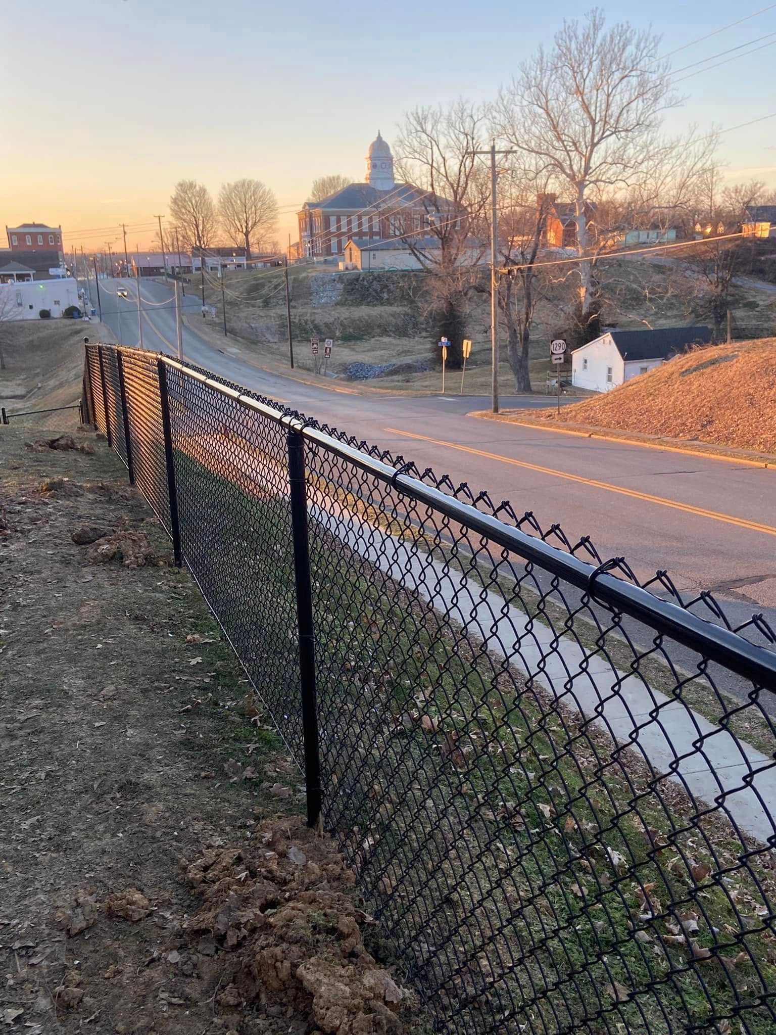 Black chain-link fence bordering a grassy hill, overlooking a road and buildings at dusk.