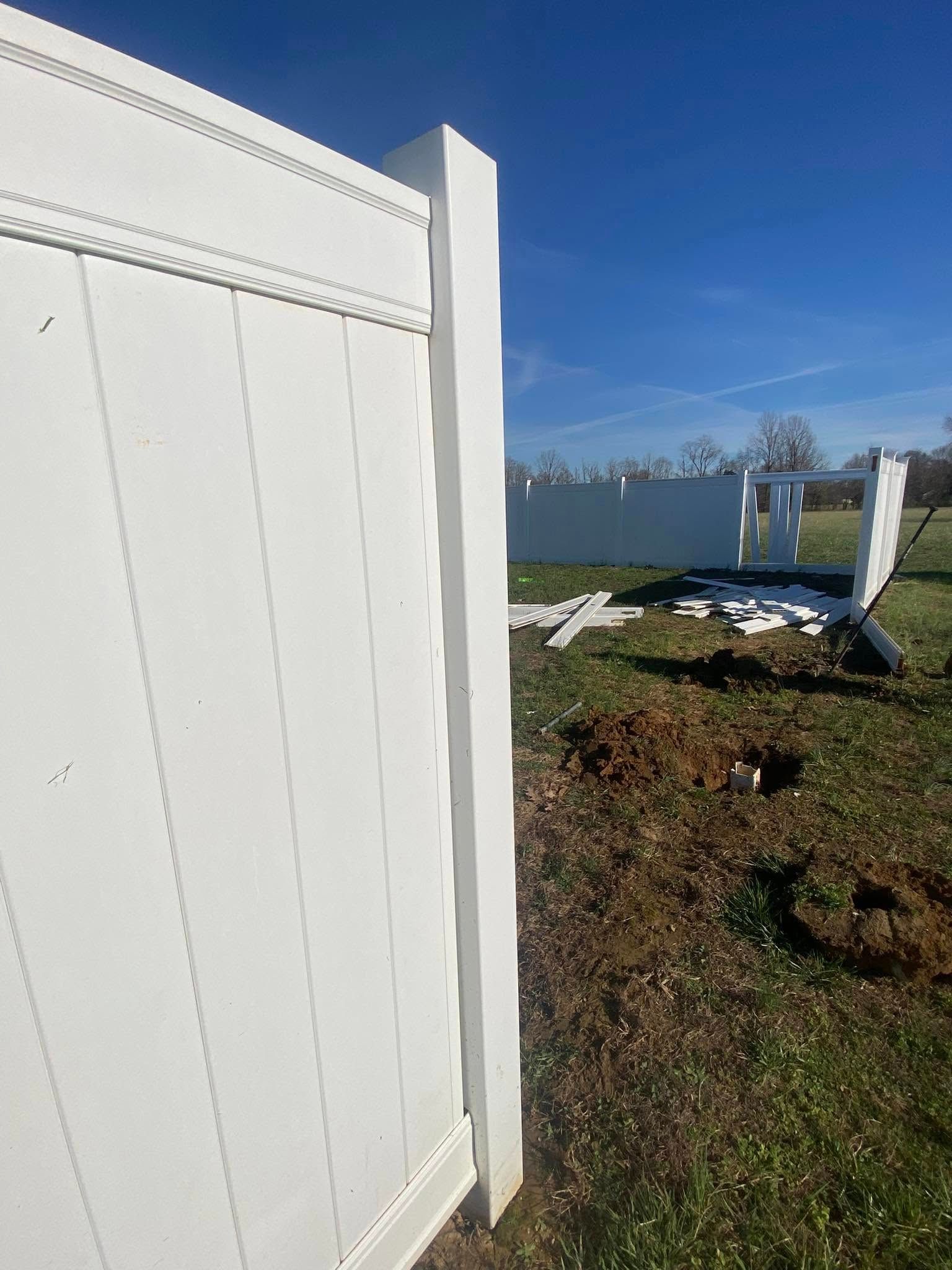 White vinyl fence under construction, posts, panels, and parts on grassy lawn, clear blue sky.