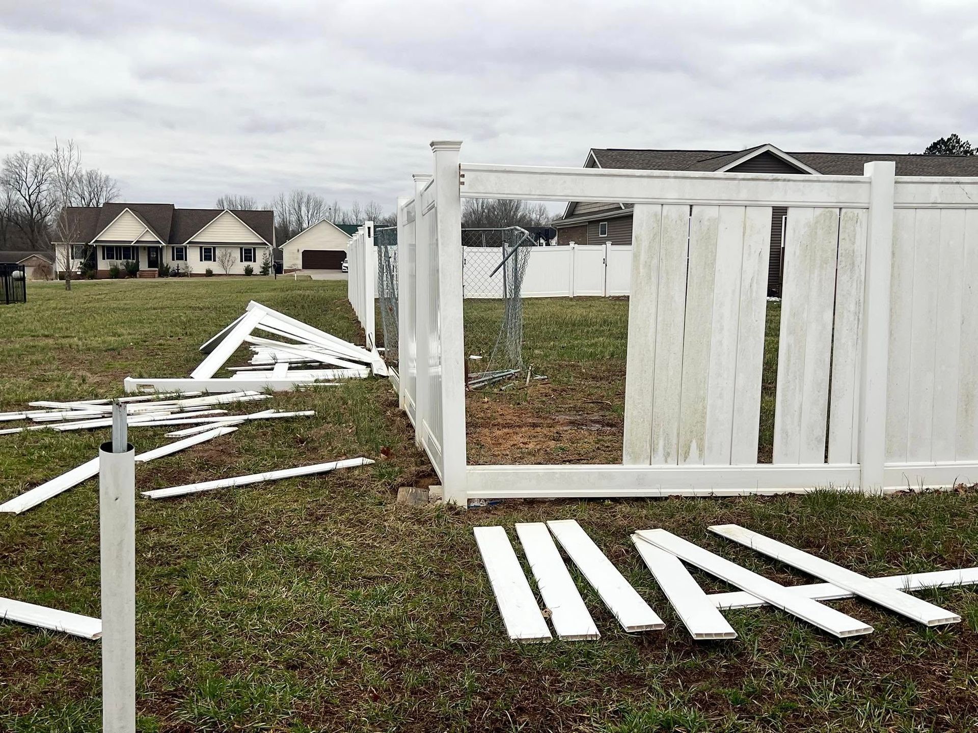 Damaged white vinyl fence with scattered pieces in a grassy yard, houses in the background under a cloudy sky.