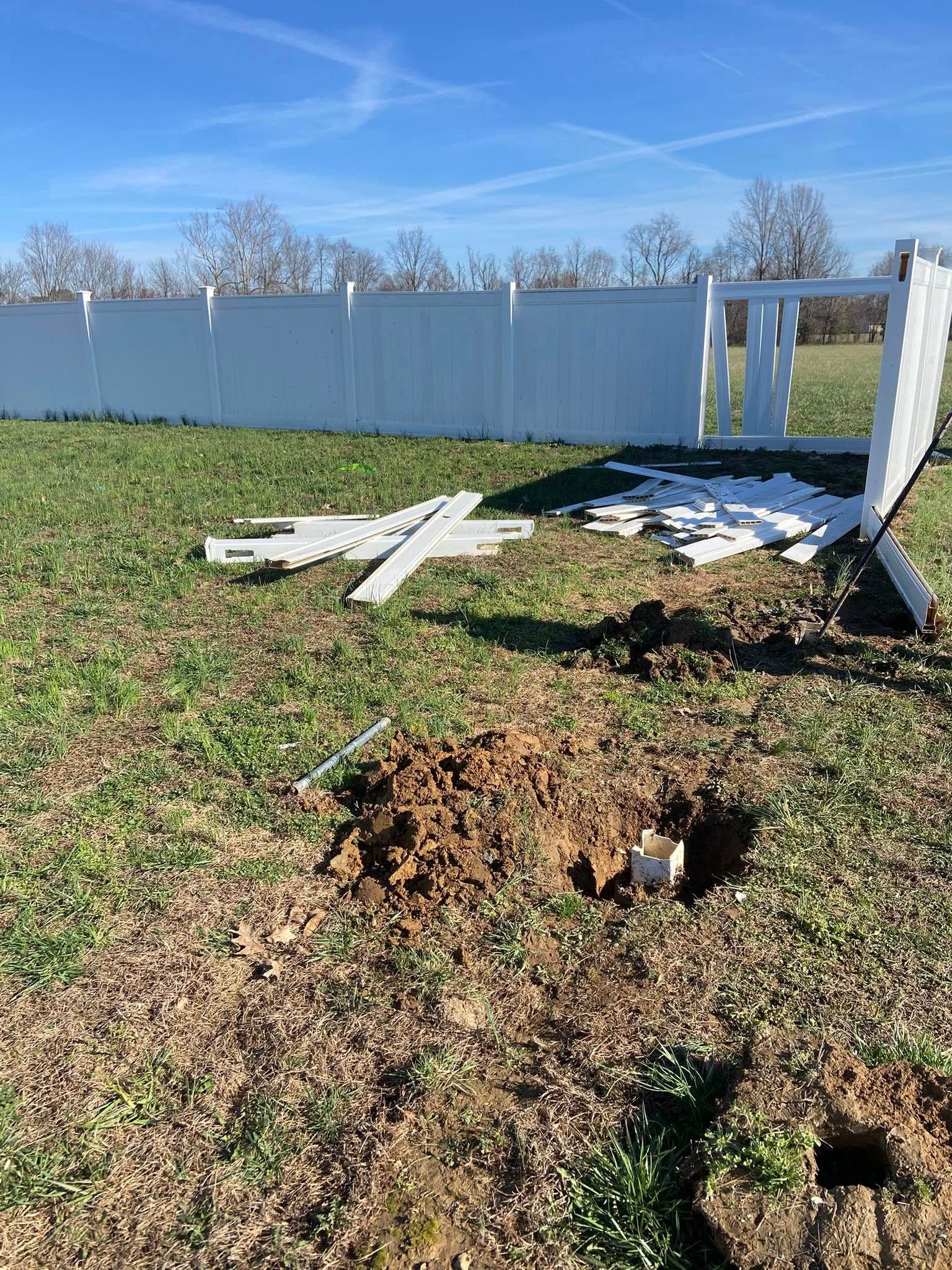 A damaged white vinyl fence with scattered pieces, a disturbed patch of earth, and blue sky in the background.