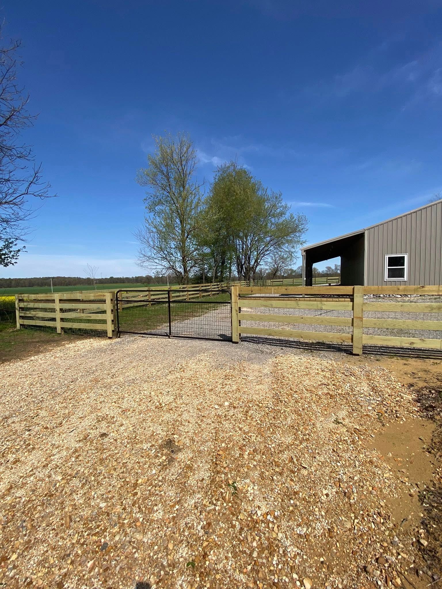 Gravel driveway leading to a gate and wooden fence surrounding a barn on a sunny day.