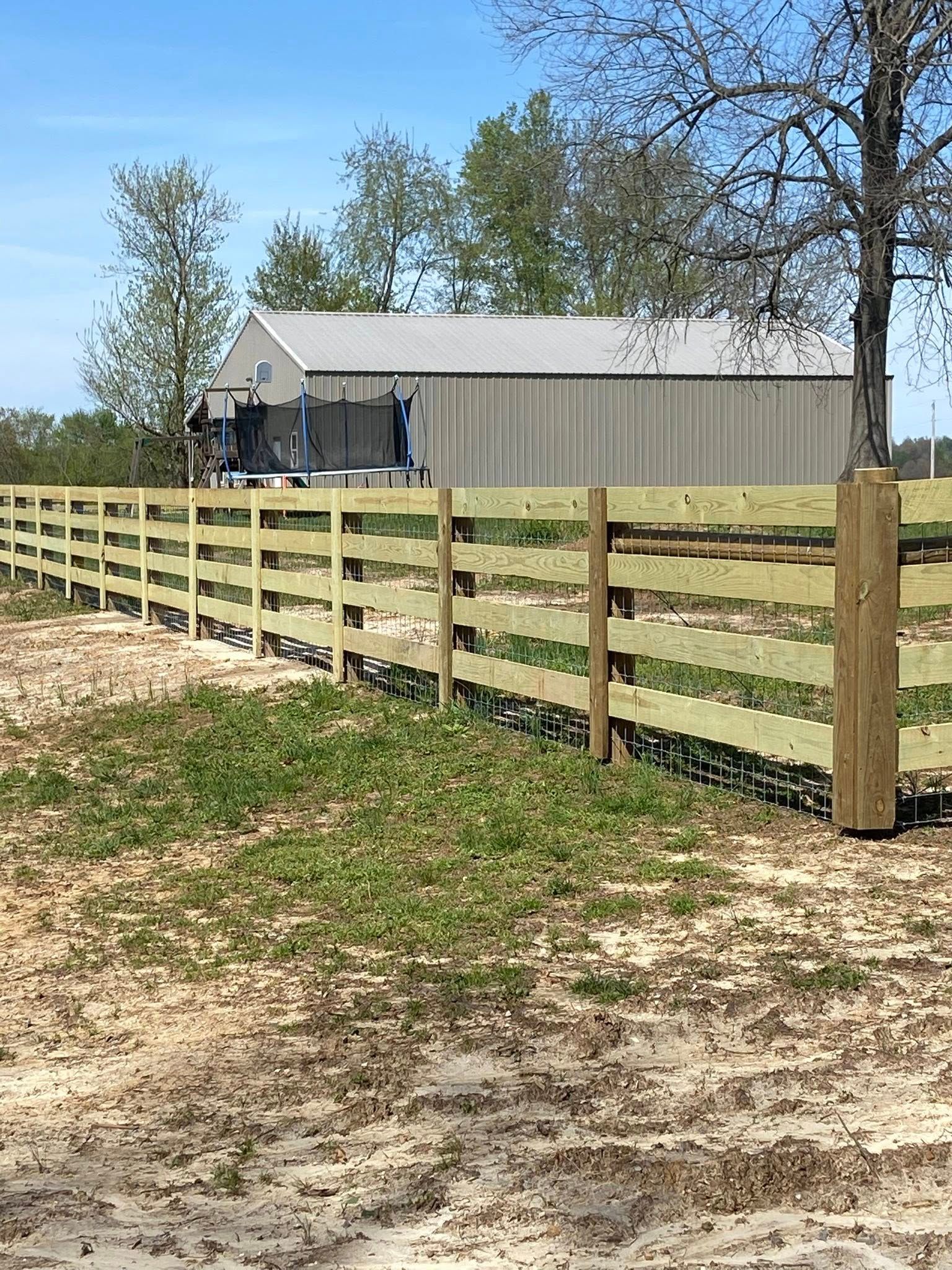 Wooden fence in front of a tan metal building with trees in the background under a blue sky.