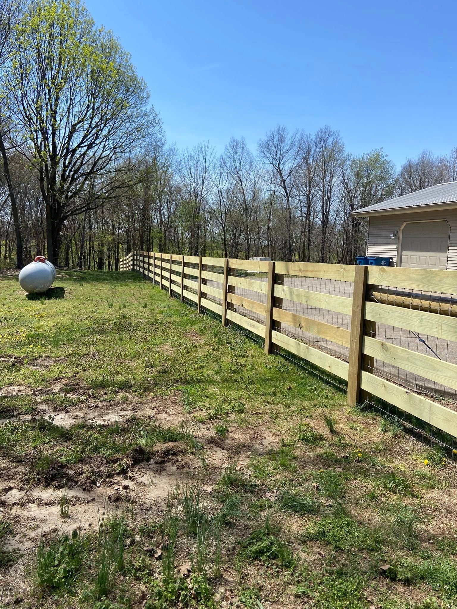 A wooden fence stretches across a grassy yard with trees and a building in the background.