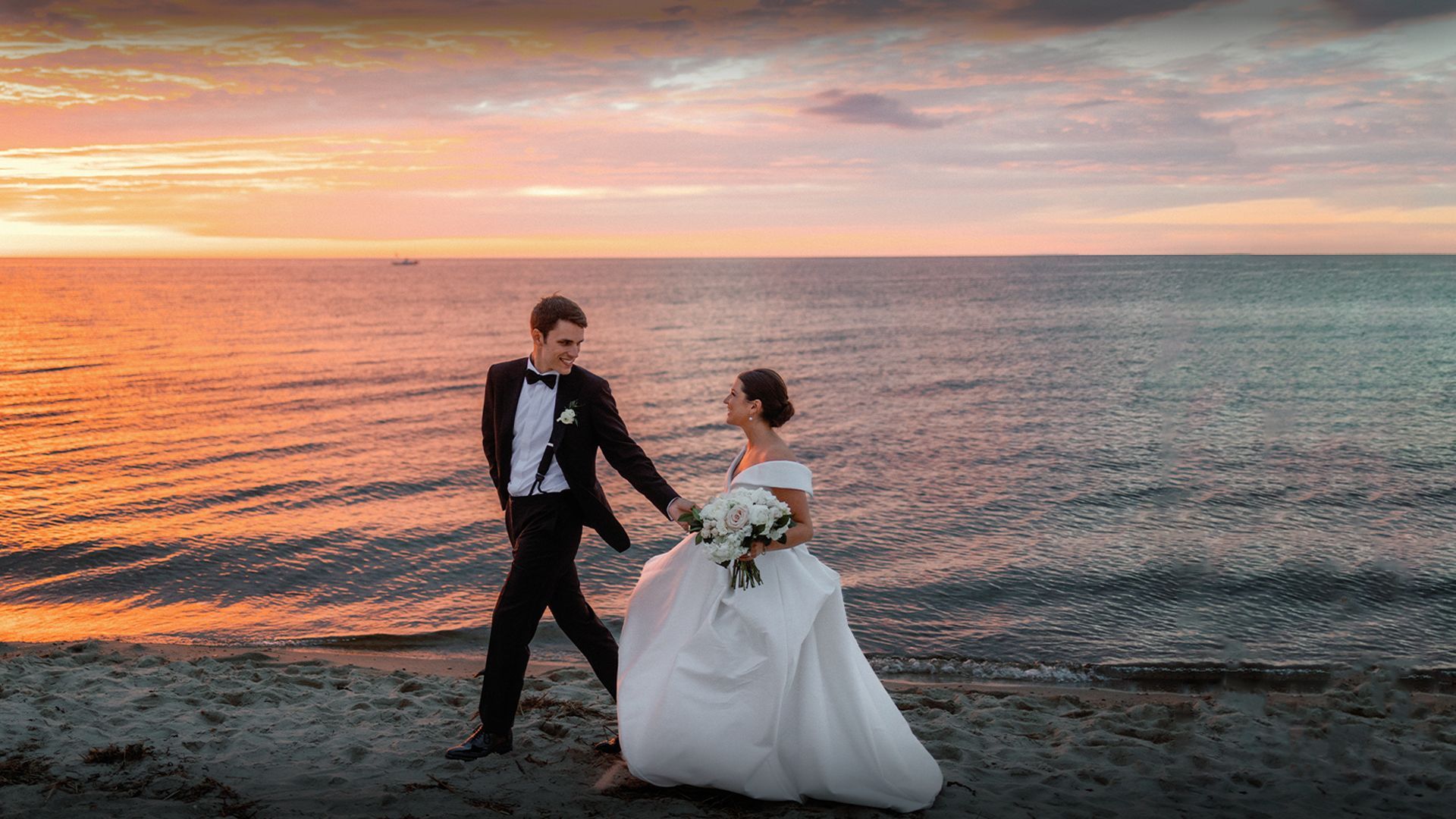 A bride and groom are walking on the beach at sunset.