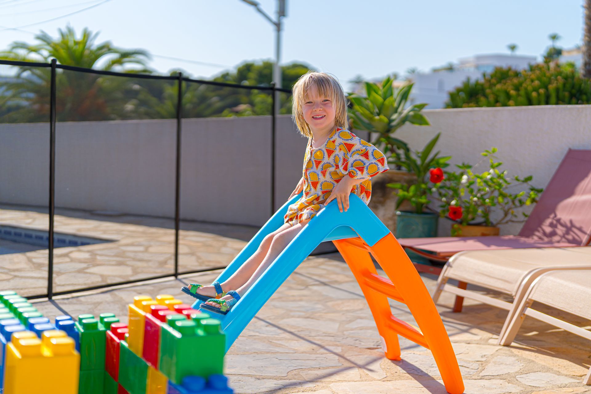 Een persoon in oranje en blauw speelt met gigantische, kleurrijke blokken naast een zwembad op een zonnig terras.