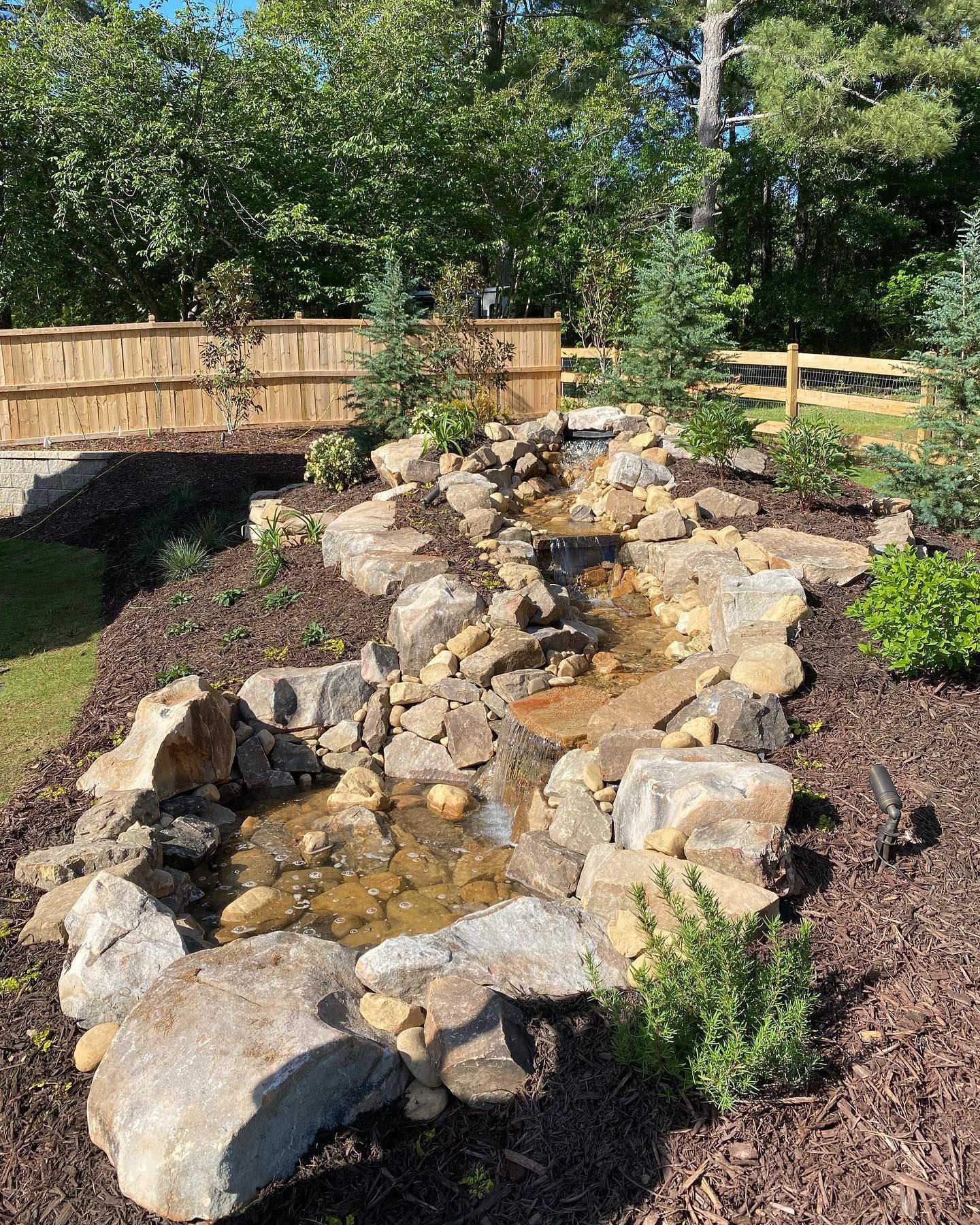 Small waterfall feature with rocks, plants, and a wooden bridge in a garden setting.