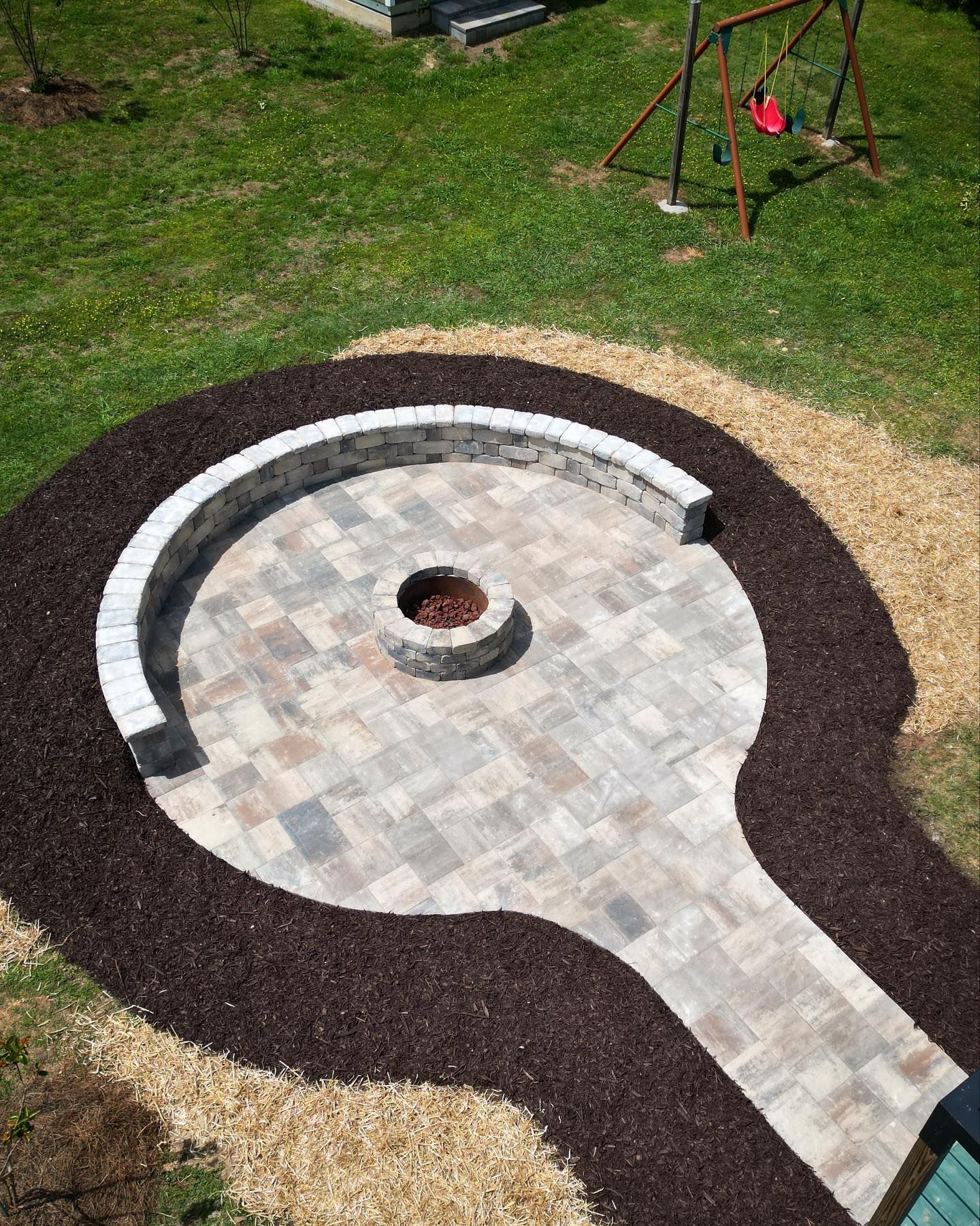 Backyard patio with fire pit, curved seating, and a pathway, surrounded by mulch and grass.