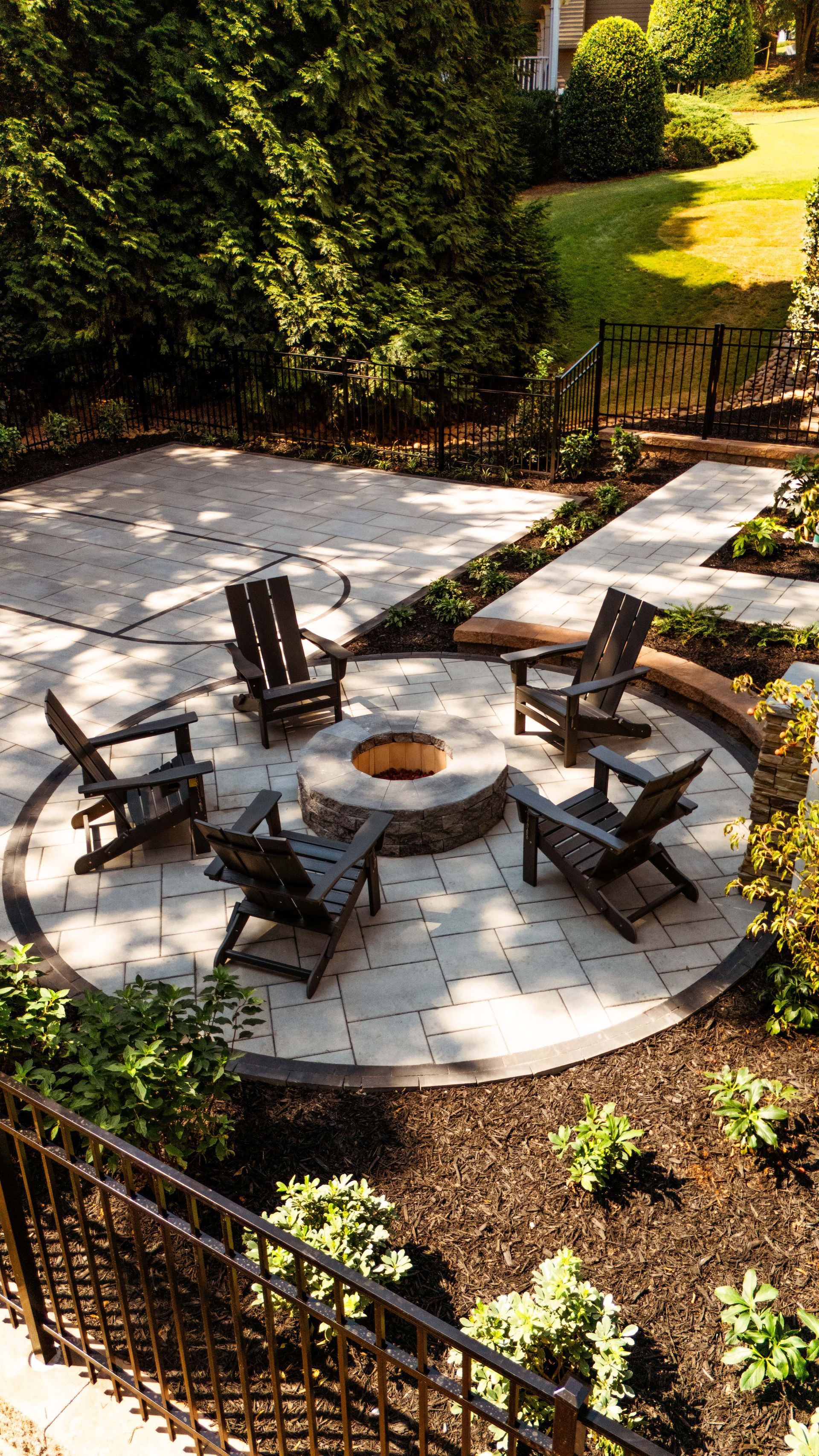 A circular stone patio with Adirondack chairs around a fire pit in a landscaped backyard.