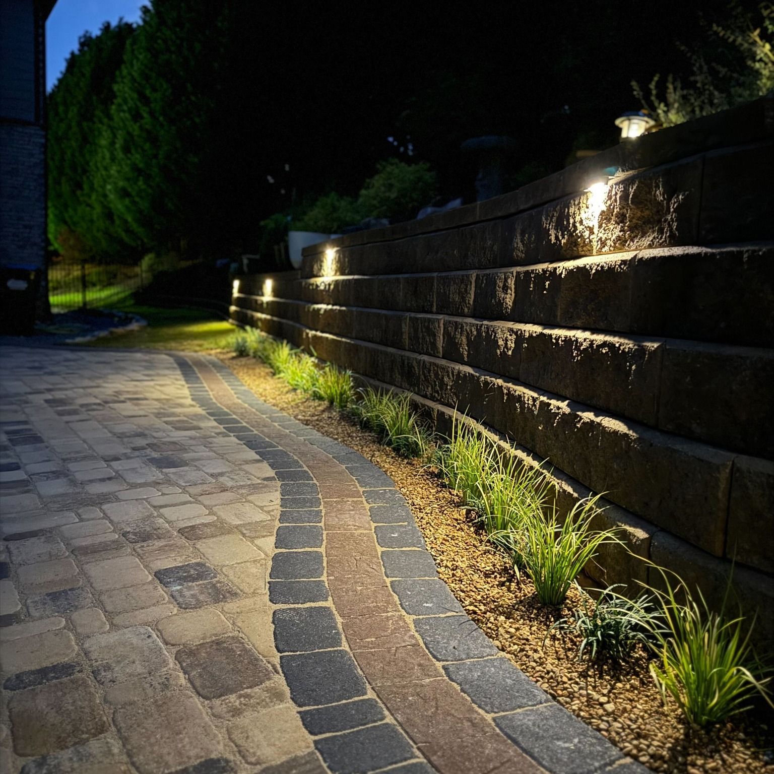 Brick pathway curves beside a tiered retaining wall, illuminated at night with accent lights.