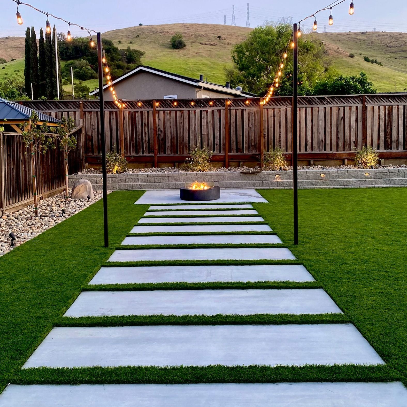 Backyard with stepping stones, fire pit, string lights, green grass, and a wooden fence.